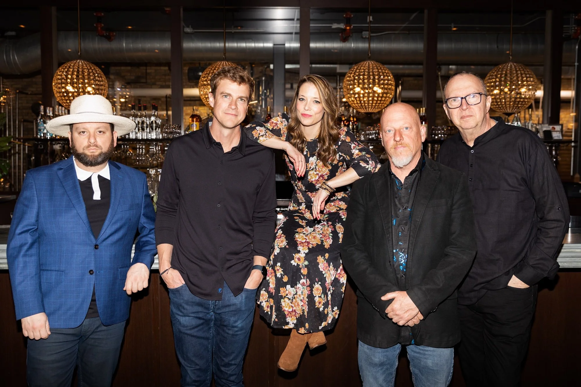 Group of five people standing in a bar or restaurant with open kitchen, three men and one woman in the center, one man on the right, all dressed in casual or semi-formal attire.