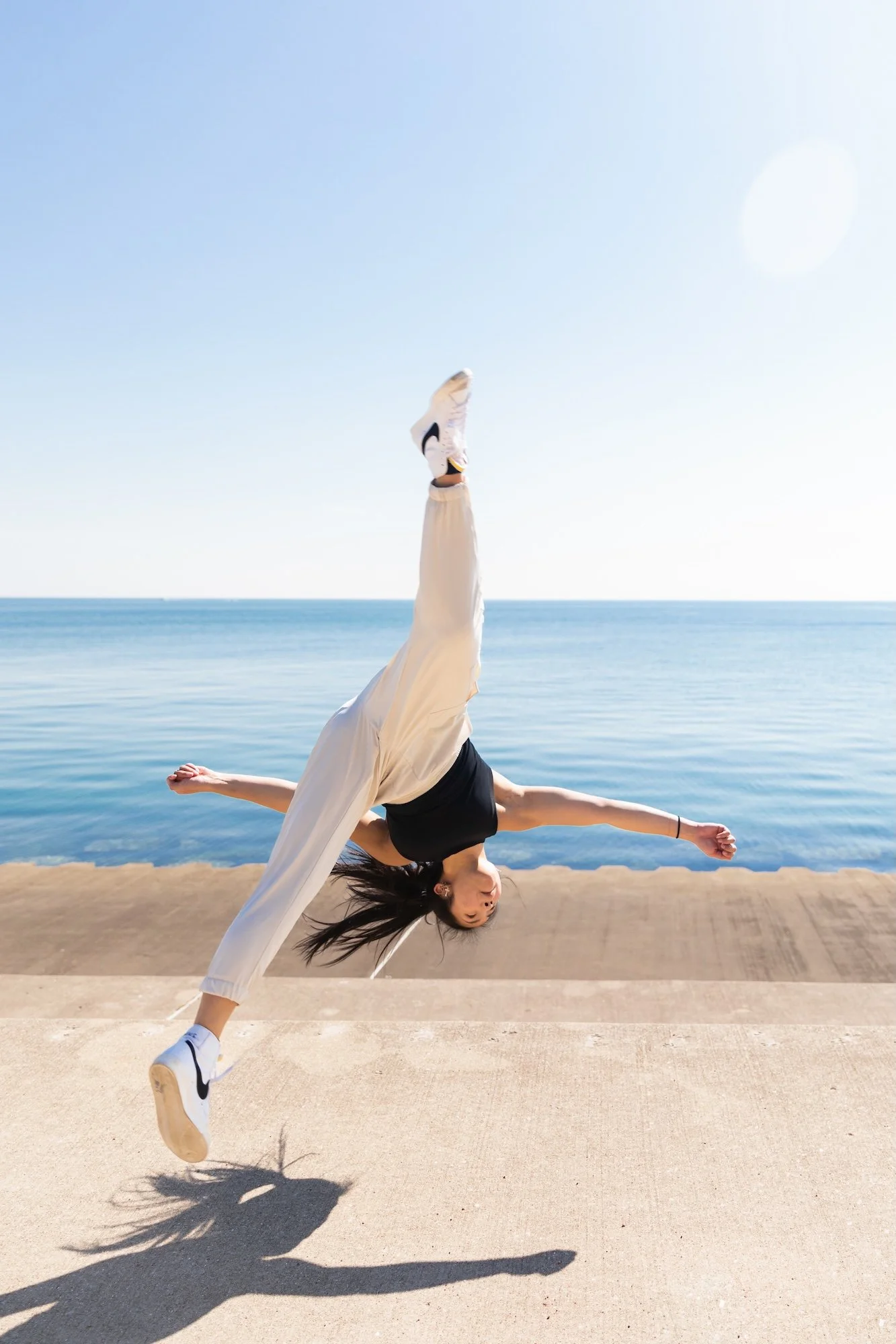 A young woman performing a breakdance move outdoors near the water, with her body upside down and one leg extended upwards, wearing athletic clothes and sneakers, on a sunny day.