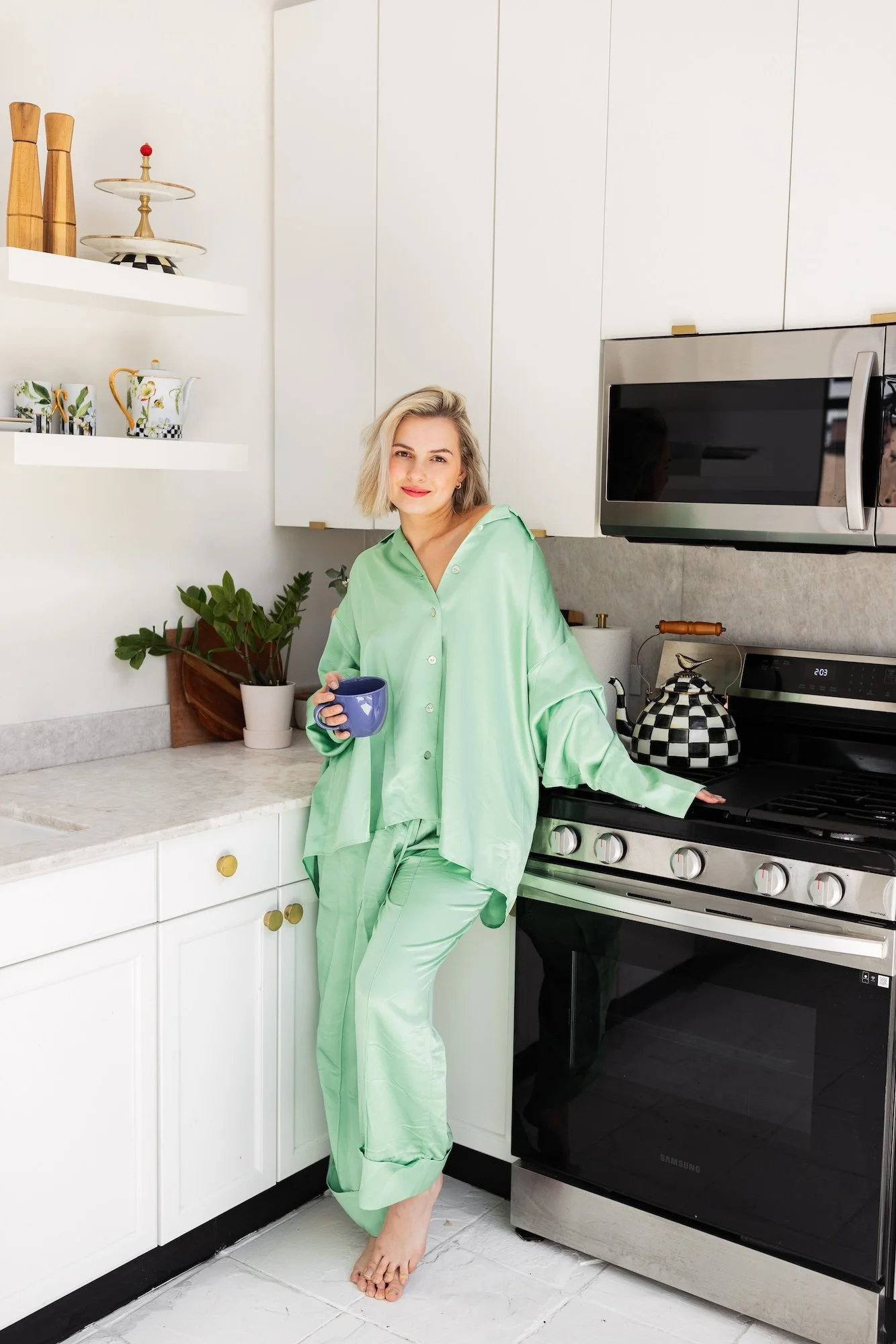 A woman in light green satin pajamas standing in a modern white kitchen, holding a purple mug, with plants, kitchenware, and appliances visible around her.