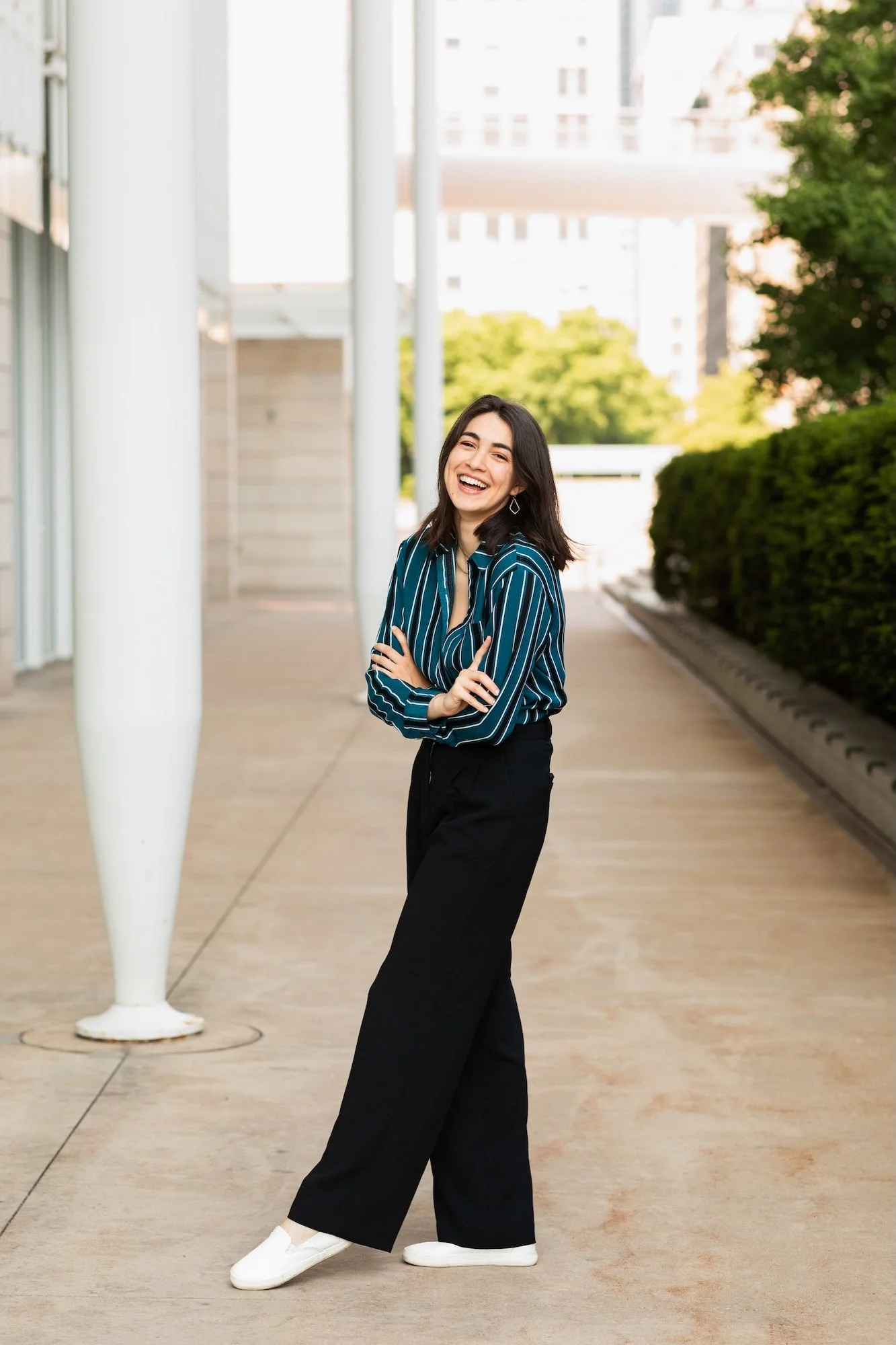 A young woman with dark hair wearing a teal striped shirt, black wide-leg pants, and white shoes, standing outdoors, smiling, with trees and buildings in the background.