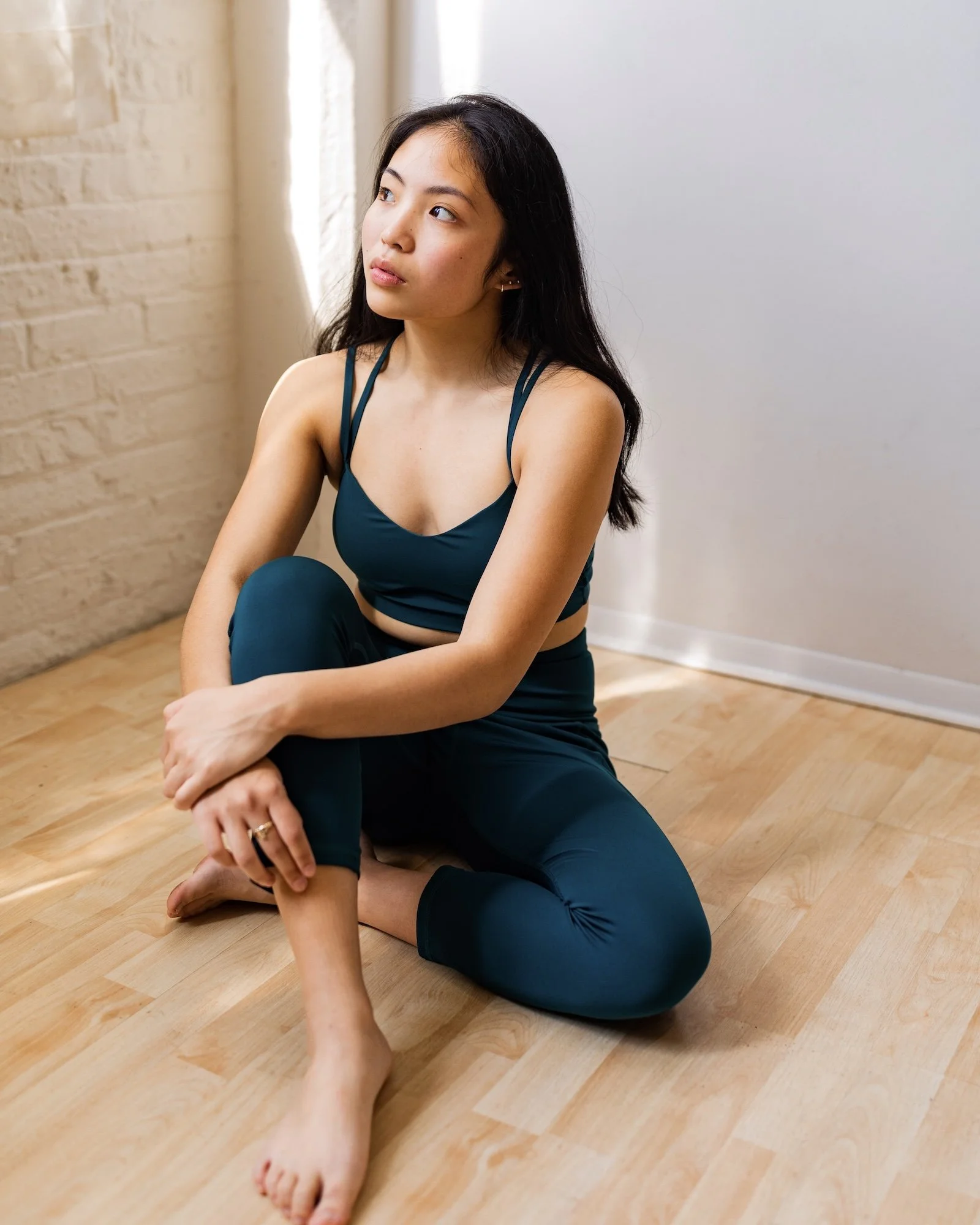 A young woman sits on a wooden floor next to a window, wearing a teal sports bra and matching leggings, with her left leg crossed over her right and her hands resting on her knee, looking thoughtful.