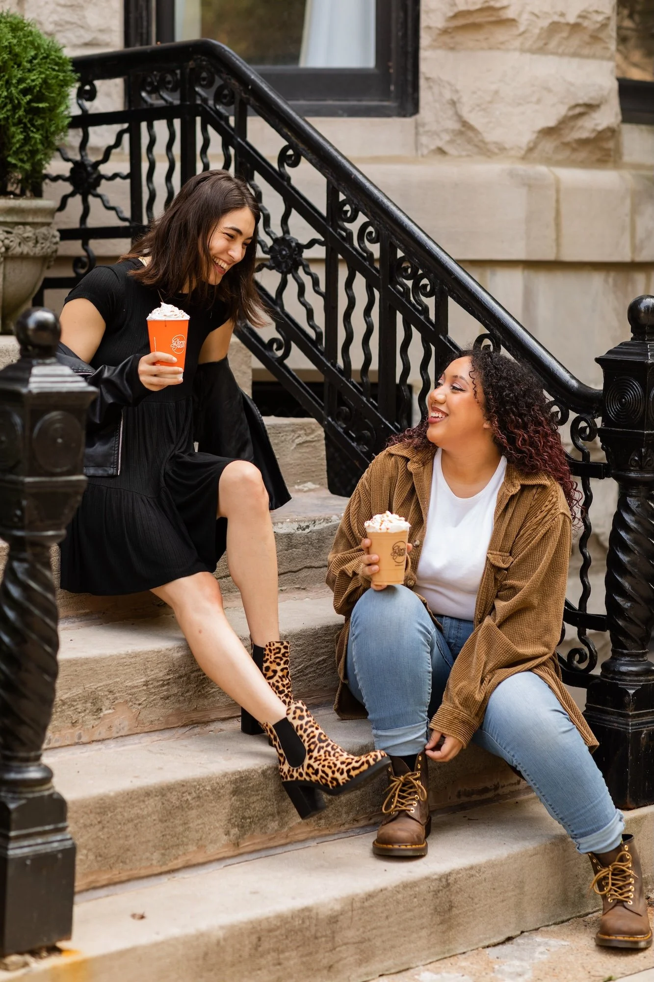 Two women chatting and smiling while sitting on steps outside. One is holding a cup of coffee, and the other is holding a cup of ice cream.