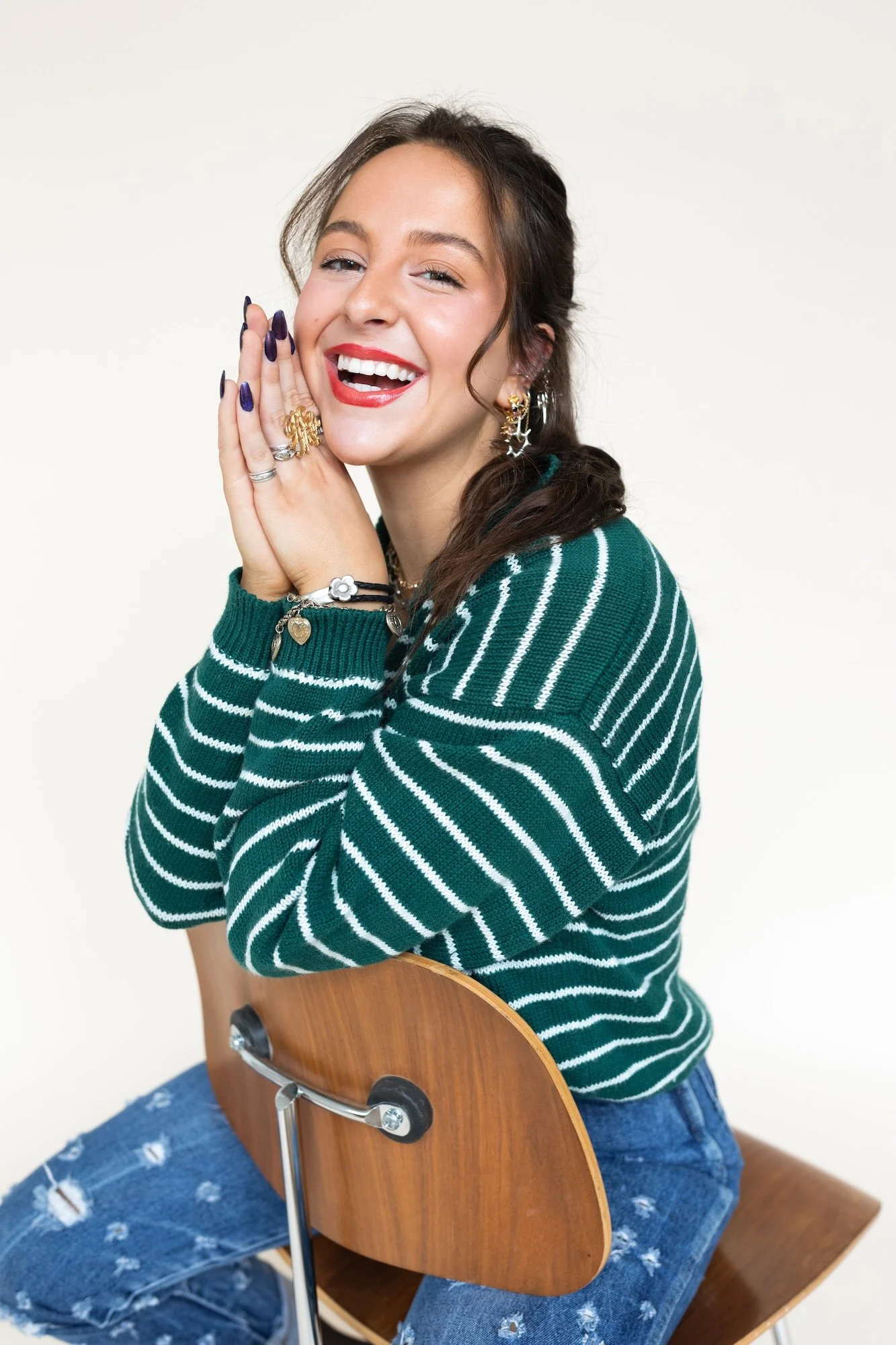 A young woman with brown hair, smiling and winking, wearing a green and white striped sweater and ripped jeans, sitting on a wooden chair with a curved backrest, against a plain light background.