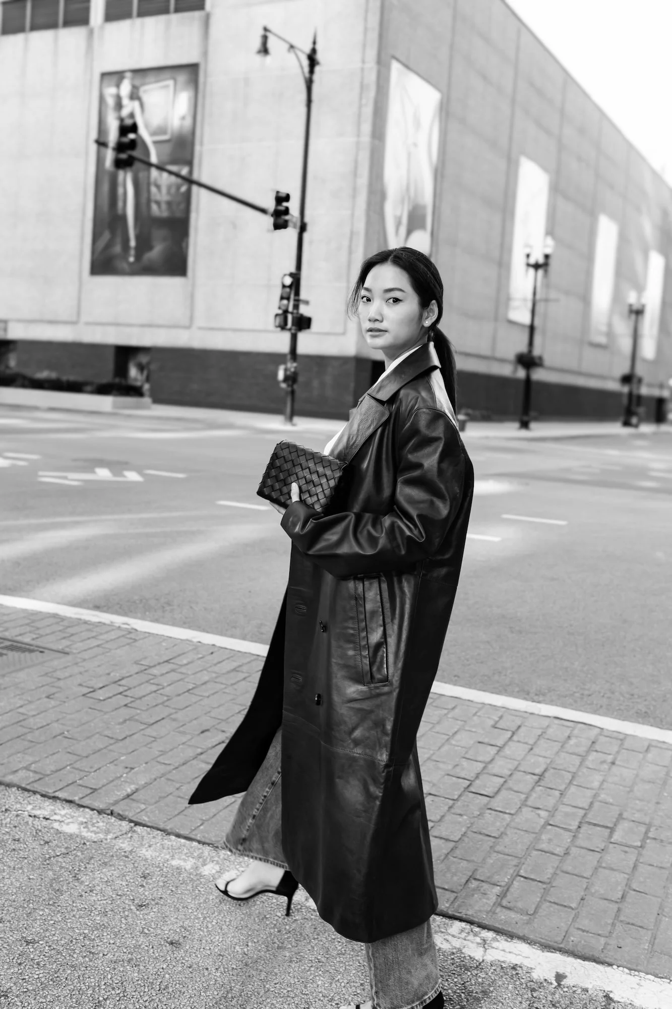 A woman standing on a city street corner holding a woven clutch purse, wearing a long leather coat and high heels, with a large modern building and traffic lights in the background.