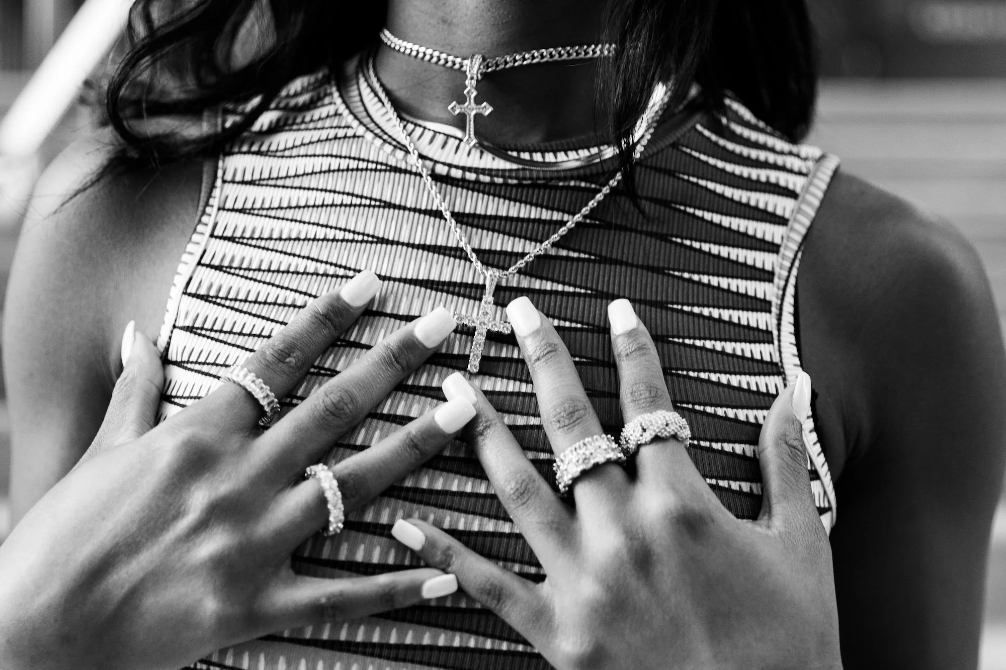 Close-up of a woman wearing layered jewelry, including multiple rings, a necklace with a cross pendant, and a beaded choker. She is touching her chest with both hands, showcasing her jewelry against her striped sleeveless top.