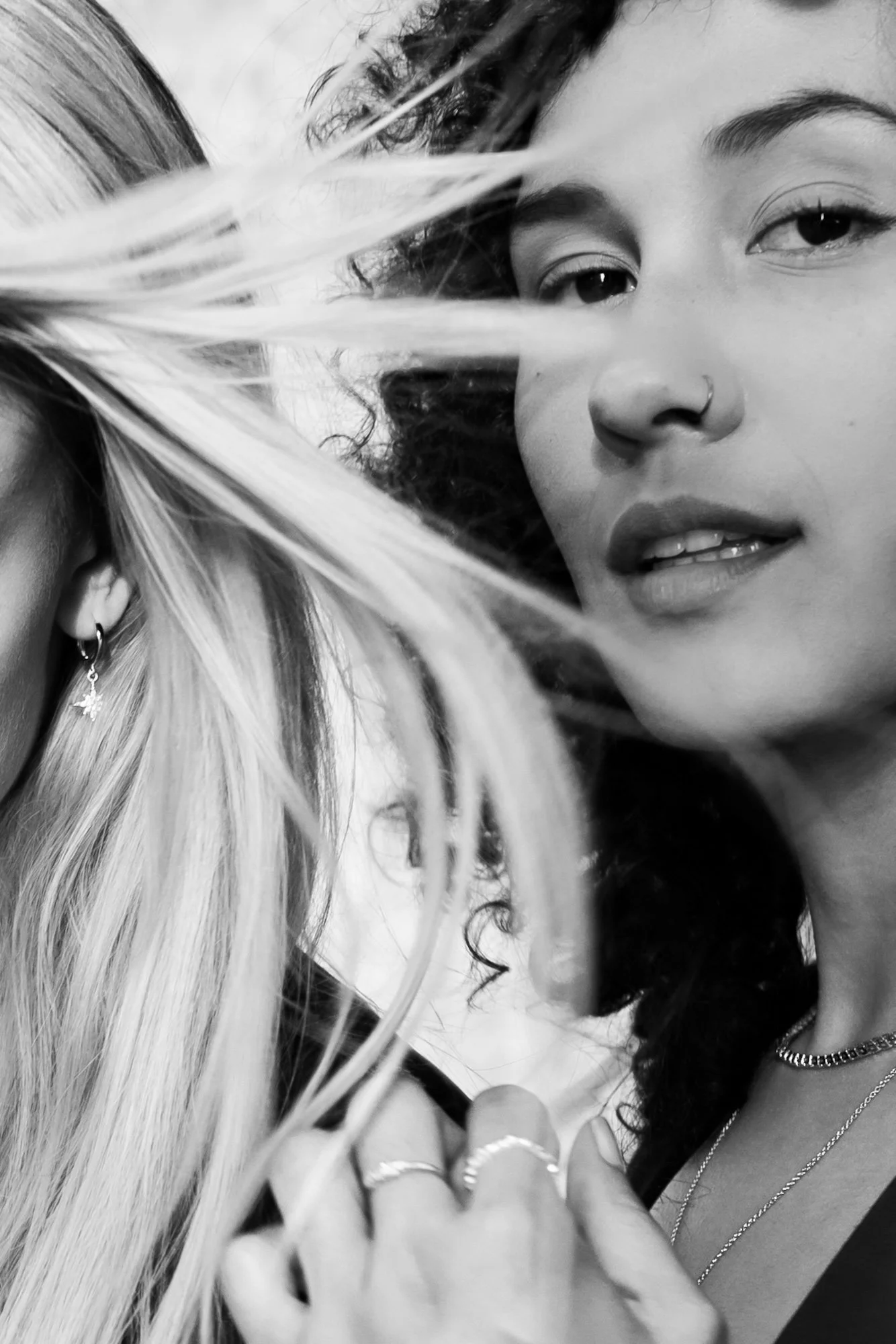 Close-up black-and-white photo of two women, with only part of the woman on the left visible. The woman on the right has wavy hair, a nose ring, light eye makeup, and is wearing rings, a chain necklace, and earrings. There are blurred plant leaves crossing in front of the subjects.