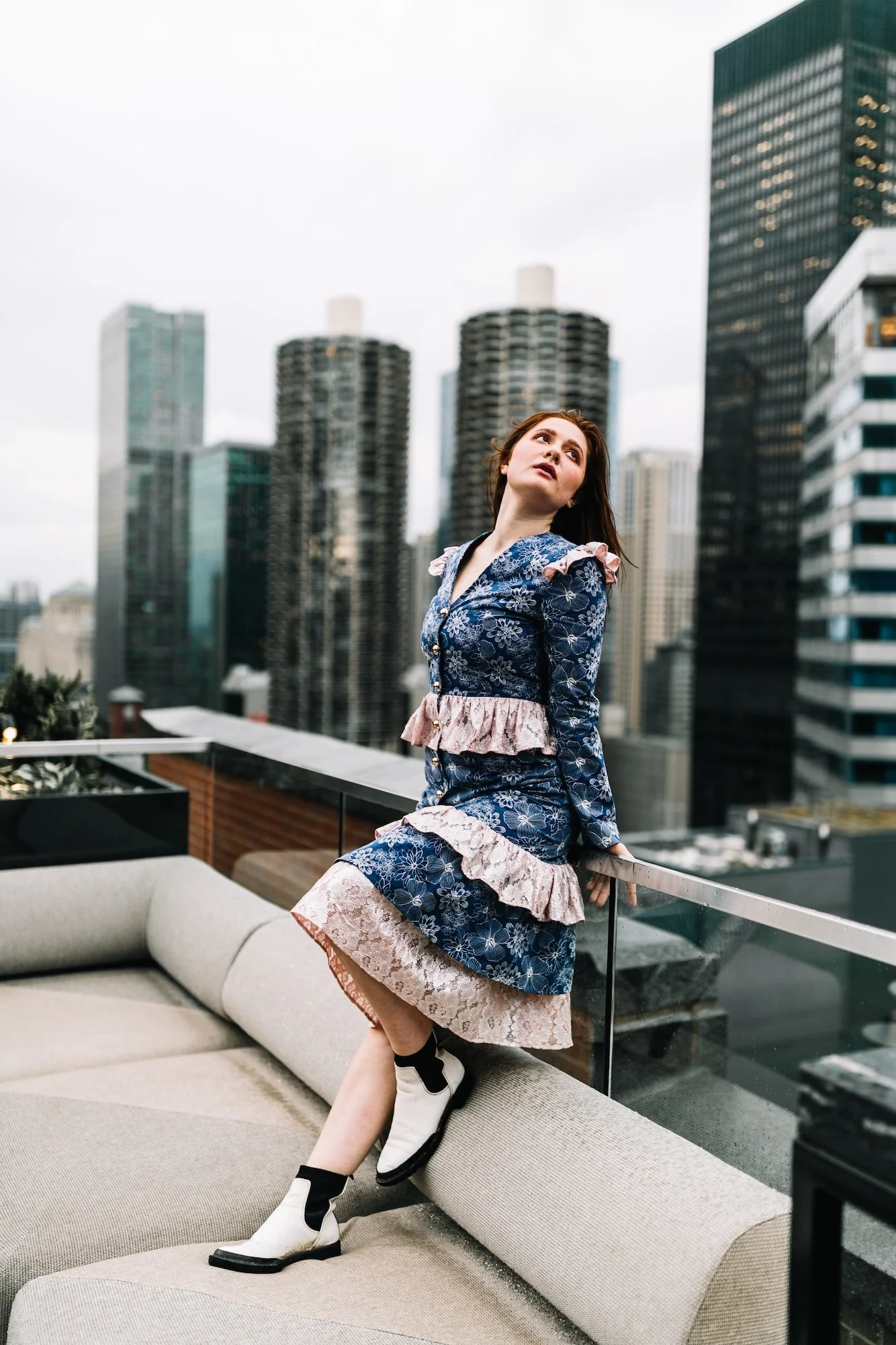 A woman in a blue floral dress with pink ruffles, standing on a rooftop with a city skyline in the background, looking upwards.