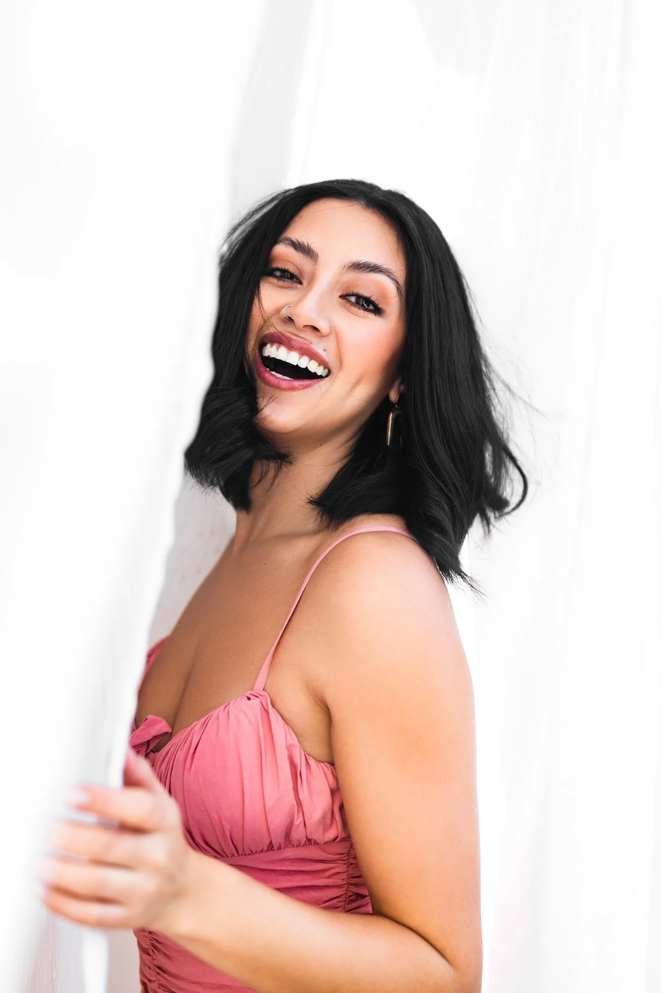 A young woman with black hair, wearing a pink spaghetti strap dress, smiling and peeking from behind a white curtain.