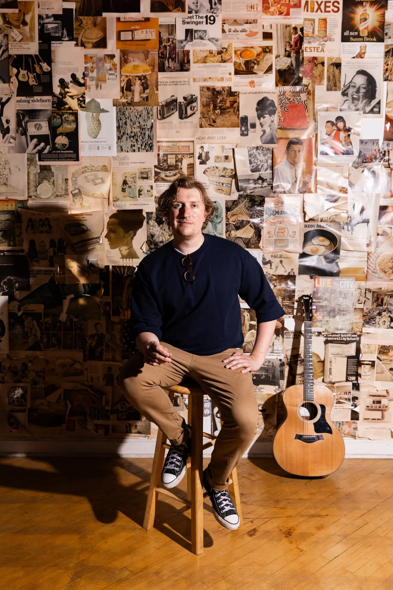 A man sitting on a wooden stool in front of a collage wall made of magazines and newspapers with a guitar leaning against the wall to his right.