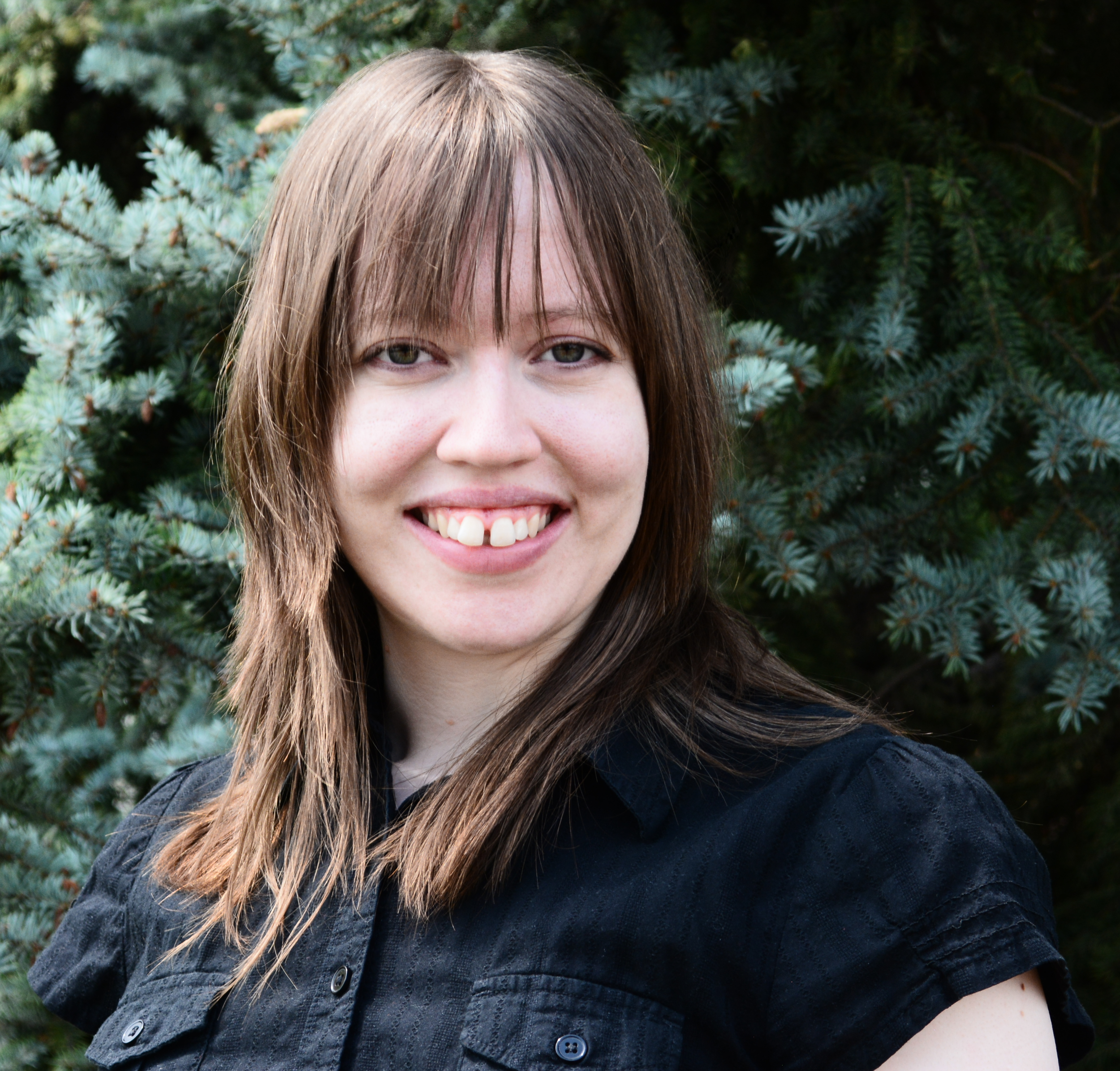 A young woman with shoulder-length brown hair and bangs, smiling, standing outdoors in front of a green pine tree. She is wearing a black short-sleeved shirt.