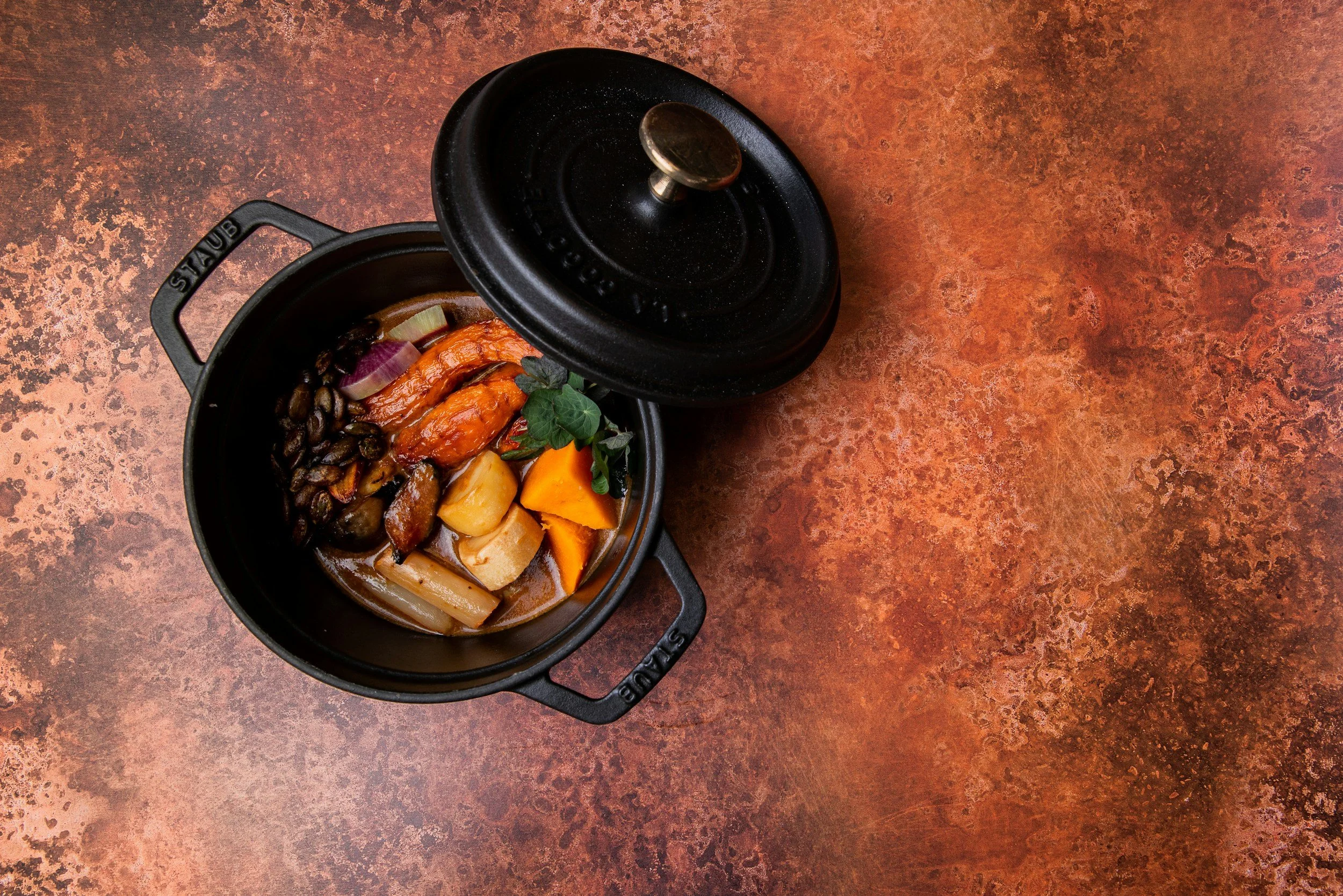 A black Staub enameled cast iron pot with the lid slightly open, containing a baked dish with roasted vegetables, beans, and herbs on a rust-colored surface.