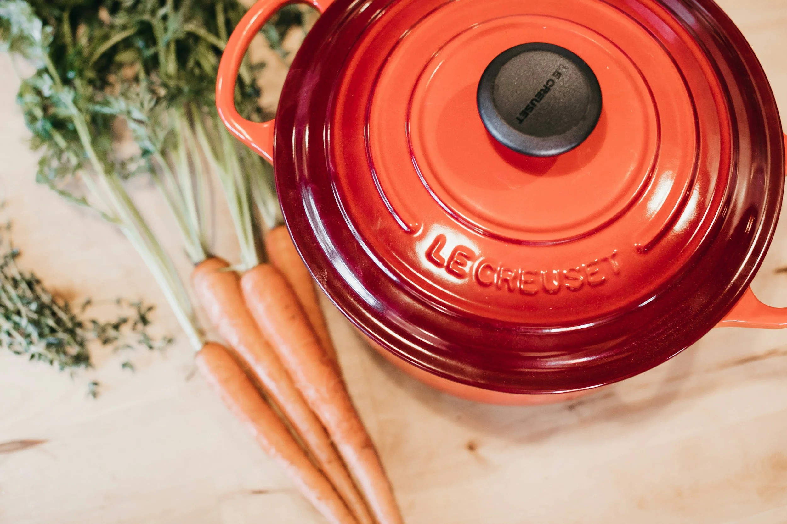 Top view of a red Le Creuset cast-iron pot on a wooden surface, with fresh carrots and a bundle of herbs nearby.