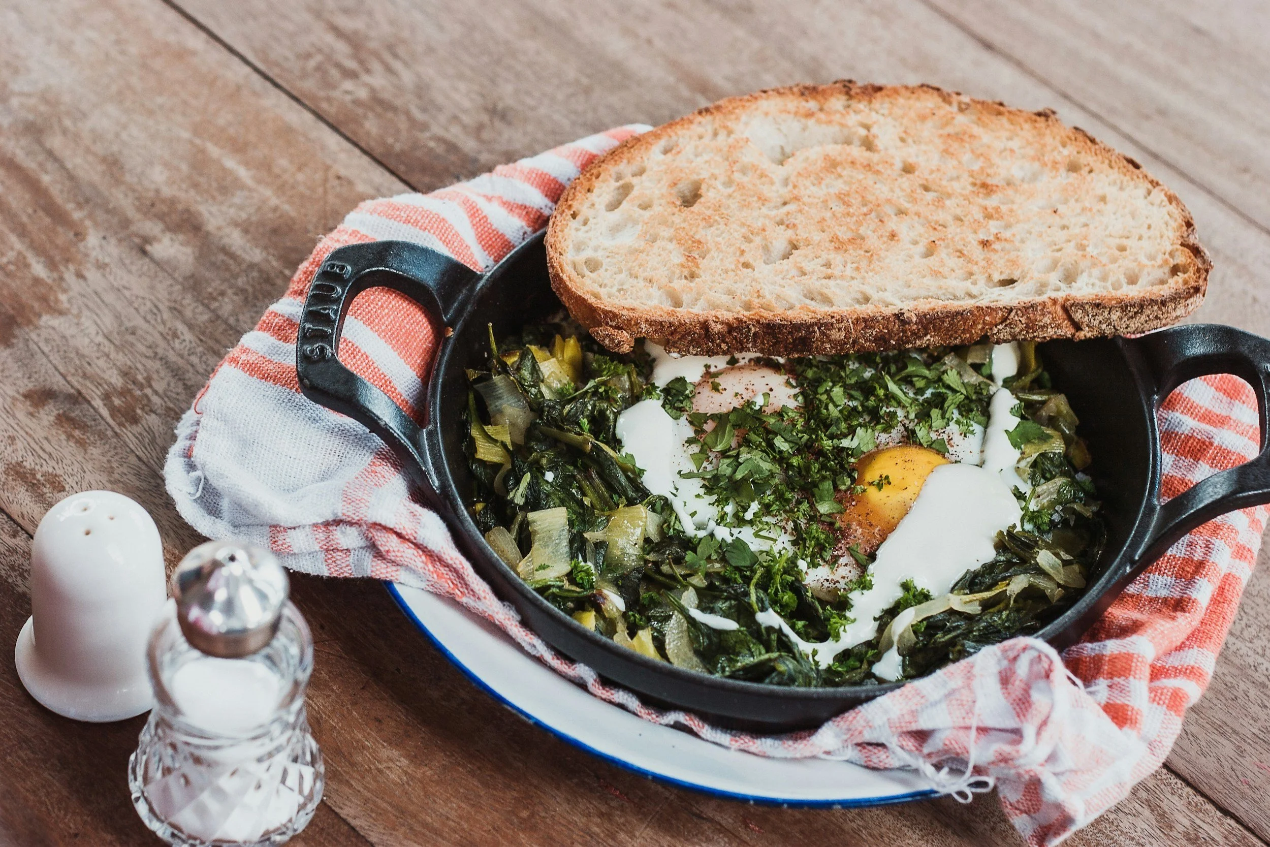 Cast iron skillet with cooked greens topped with eggs, garnished with herbs, and a slice of bread on top, resting on a striped cloth on a wooden table with salt and pepper shakers nearby.