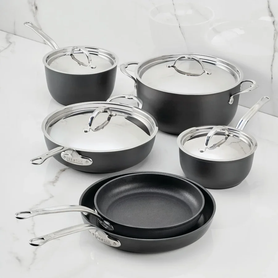Set of black and silver Hestan non-stick pots and pans with lids on a white marble countertop.