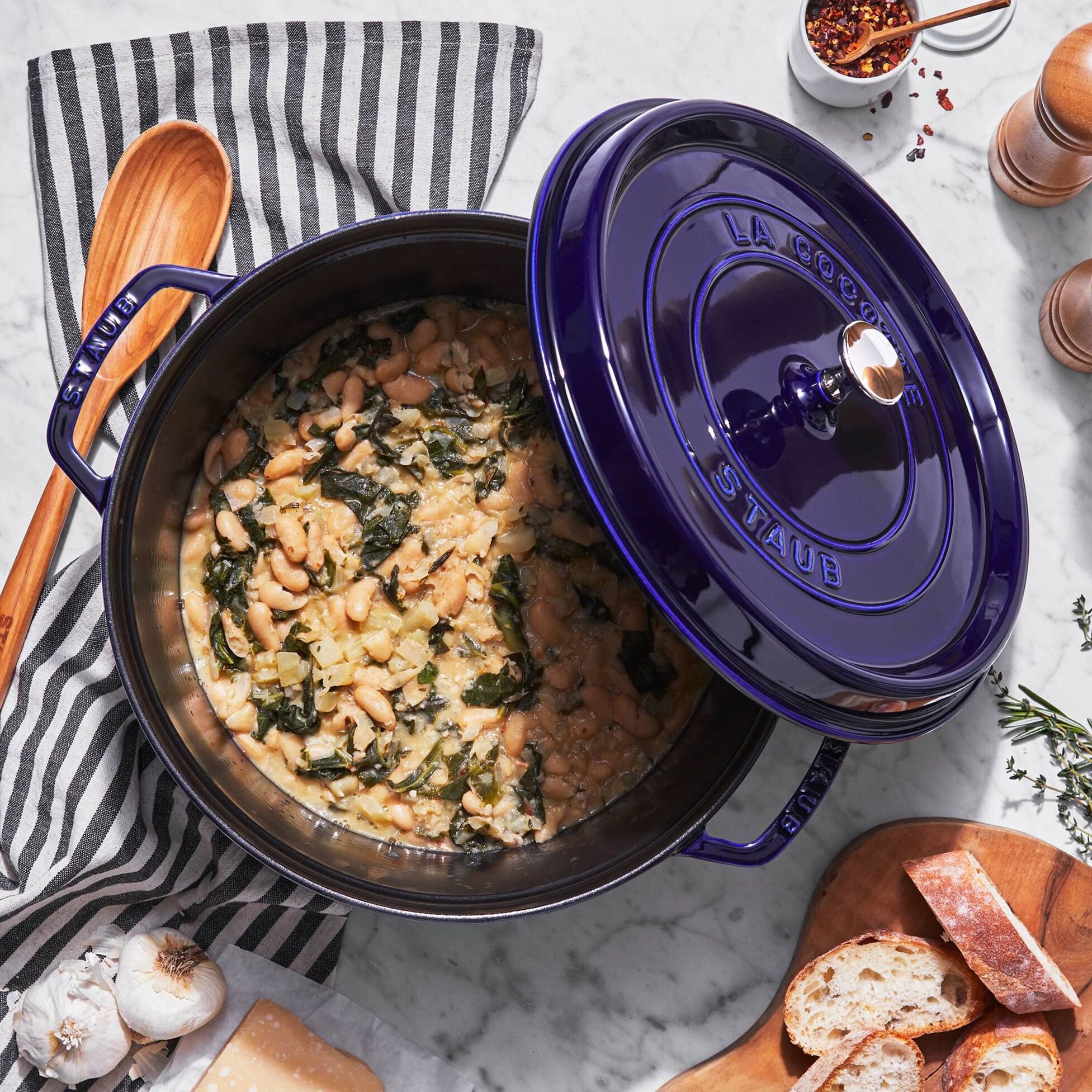 A blue Staub enameled cast iron pot filled with white bean and kale soup on a white marble countertop, with a striped cloth, garlic, bread, and spices nearby.