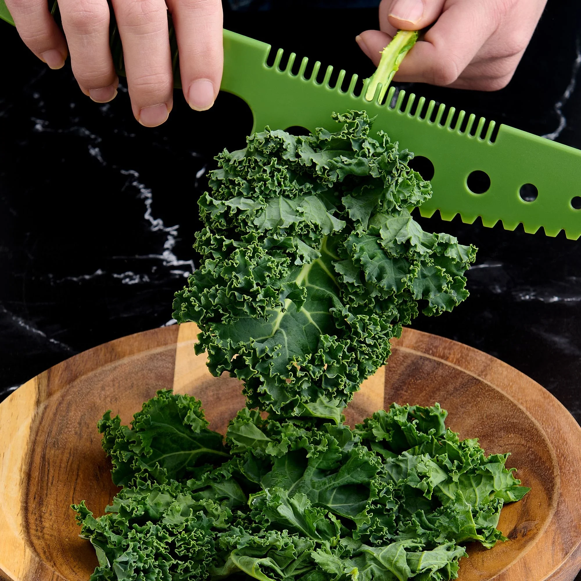 Person using a green plastic herb stripper to remove kale leaves off of the stem, over a wooden bowl.