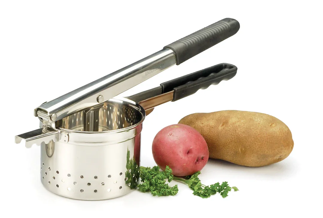 A metal potato ricer with a black handle, a raw potato, a sweet potato, and fresh parsley on a white background.