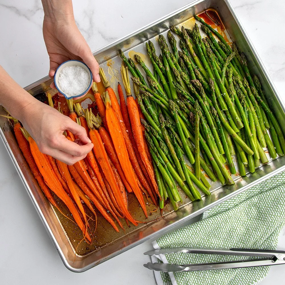 Someone sprinkling salt on roasted carrots and asparagus on a baking sheet.