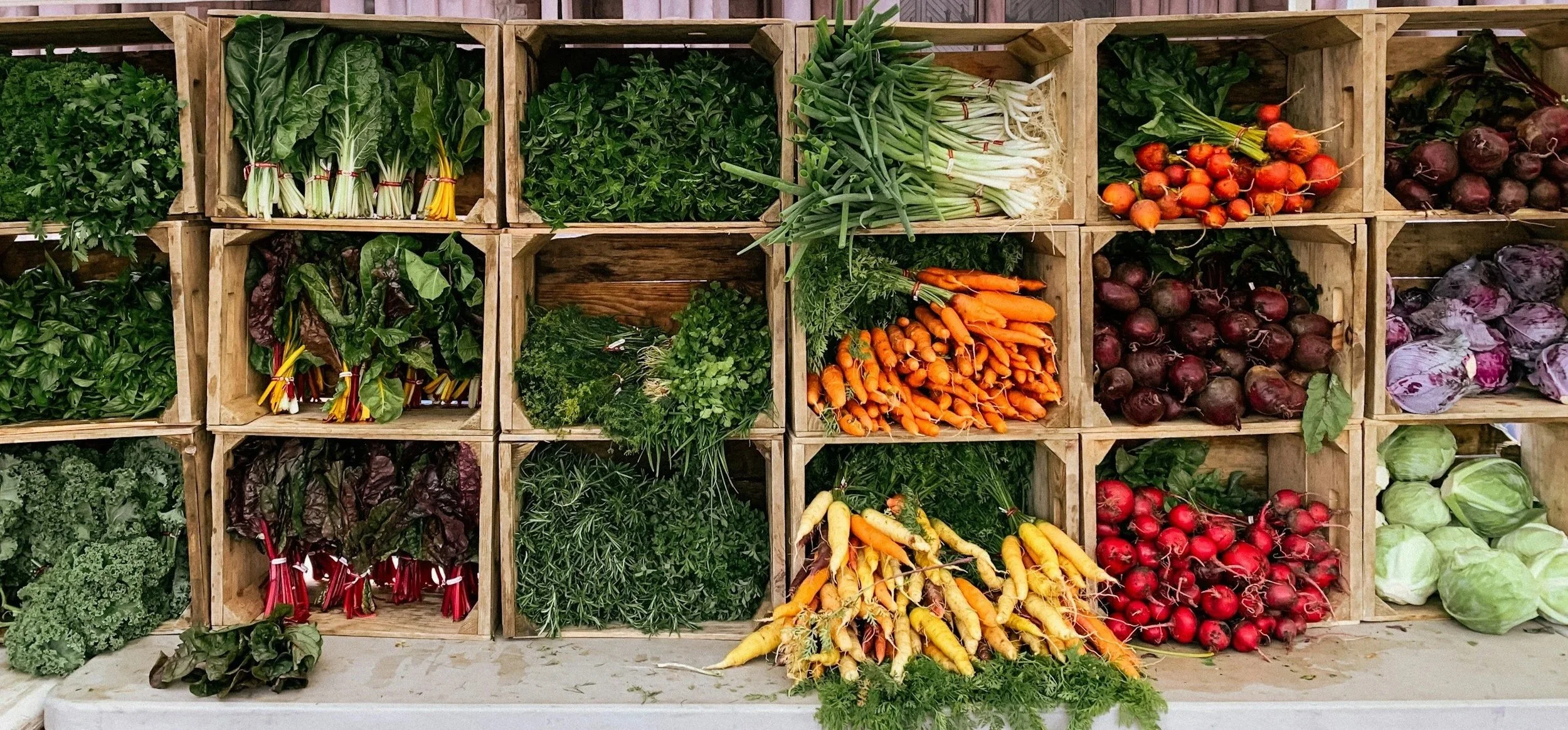 Fresh vegetables displayed in wooden crates including greens, carrots, beets, radishes, and onions.