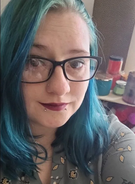Portrait of Amy, a Bakersville Fix N’ Shine team member, with teal-blue hair and black glasses.