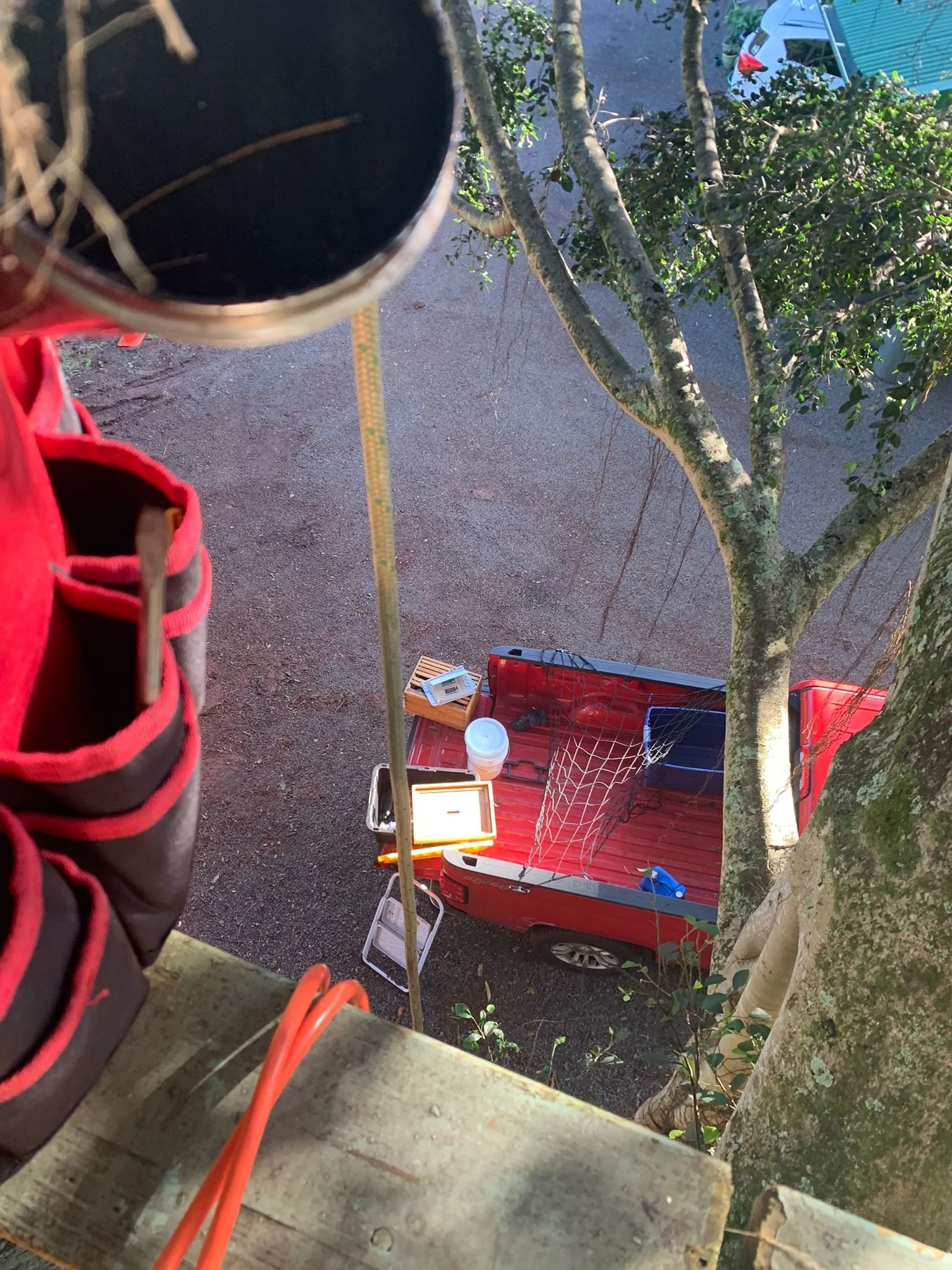 Photo taken from a tree house or high platform looking down at a red pickup truck parked on a gravel driveway. The truck's bed contains various items including a small grill, a bucket, and a blue object. There is a large tree with branches and leaves
