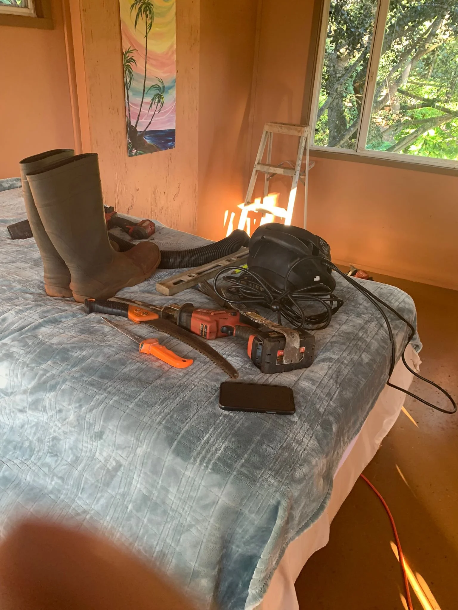 A room with a wooden interior, a large window showing green trees outside, and a bed covered with a blue quilt. On the bed are various tools including a pair of brown work boots, a vacuum hose, a power drill, a cordless saw, a smartphone, and a black