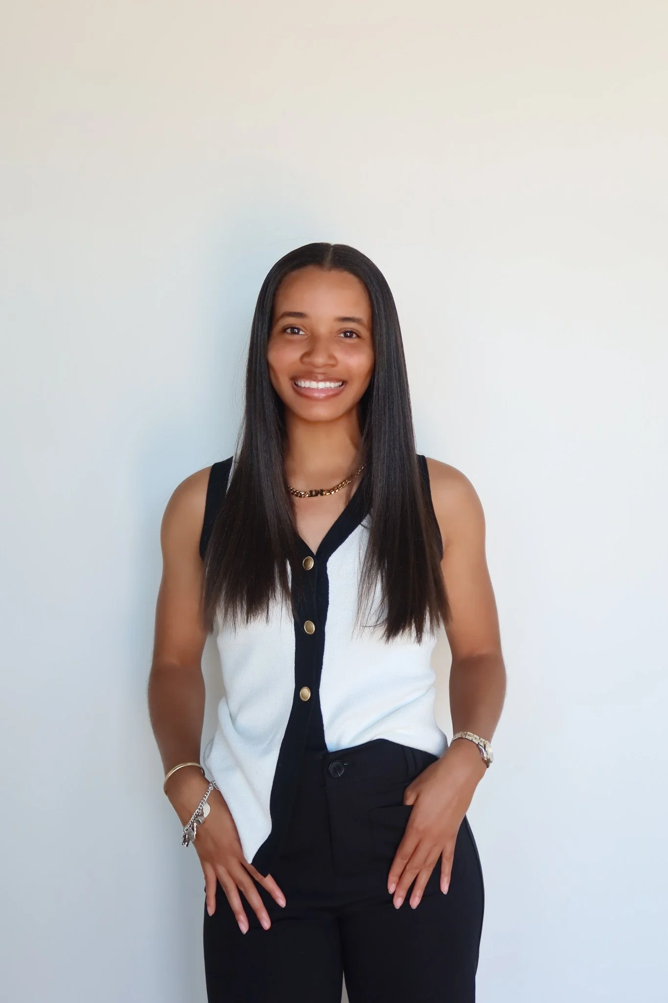 A young woman with long dark hair, smiling, wearing a sleeveless black and white button-up top, black pants, a gold chain necklace, and jewelry on her wrists, standing against a plain white wall.