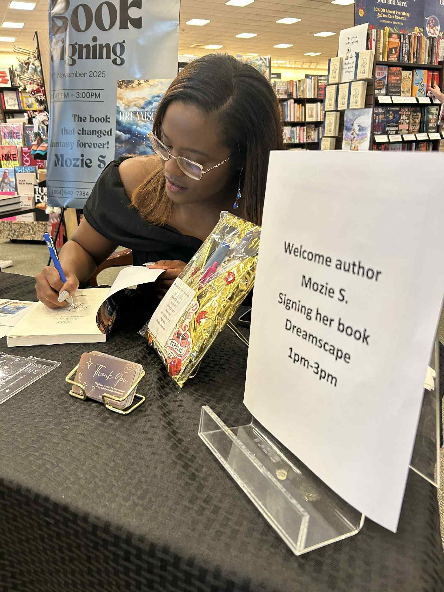 A woman signing a book at a bookstore event with a sign inviting her to sign her book, 'Dreamscape,' for fans. The event is scheduled from 1 pm to 3 pm. The woman has shoulder-length hair, glasses, and is wearing a black top. Shelves of books are visible in the background.