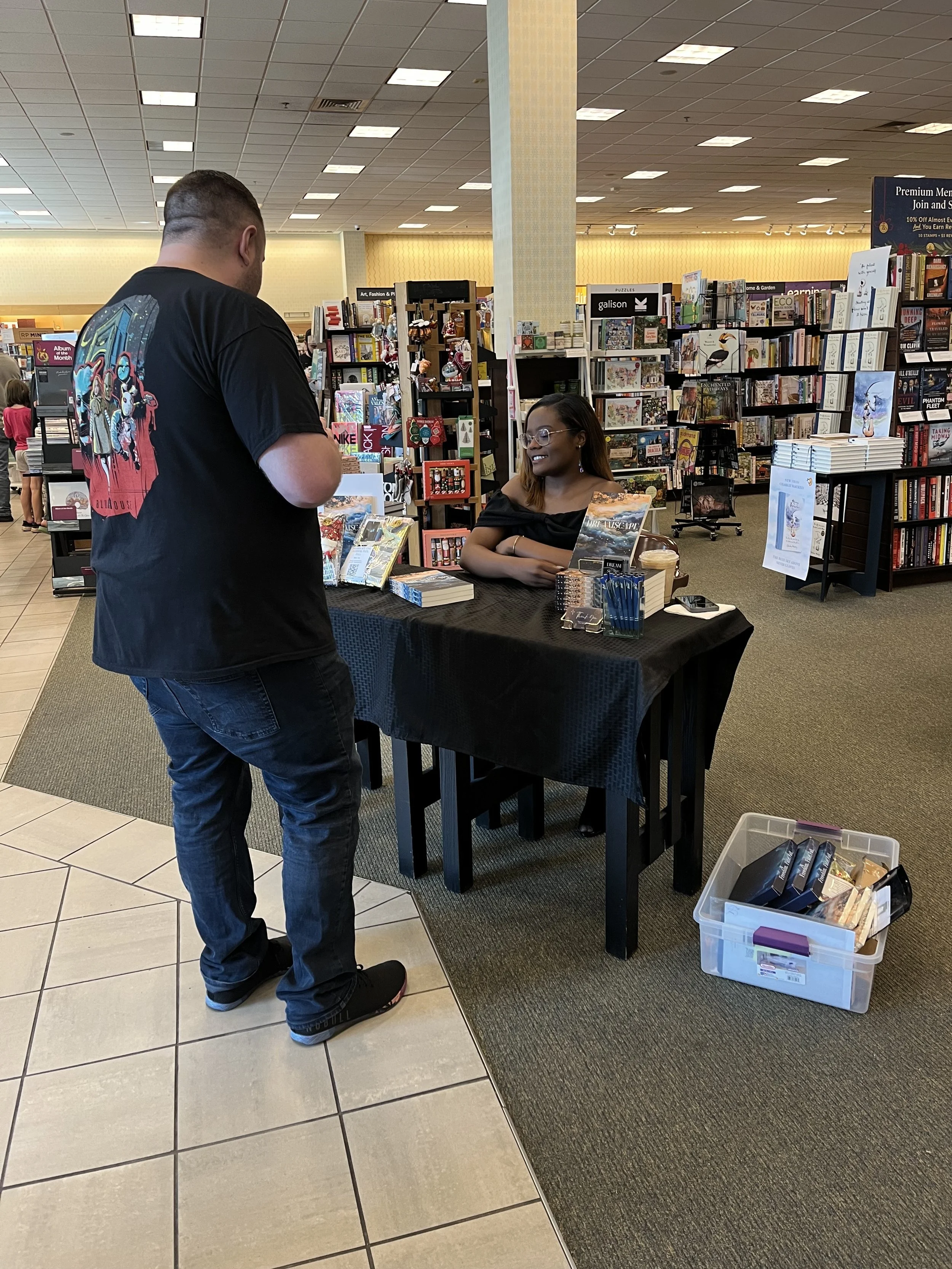 A man and a woman at a book signing event in a bookstore. The woman is sitting behind a table with books, smiling, and the man is standing in front of her, holding a book. The table has a black tablecloth, and there is a cardboard box with books on the floor nearby. Bookshelves filled with books are visible in the background.