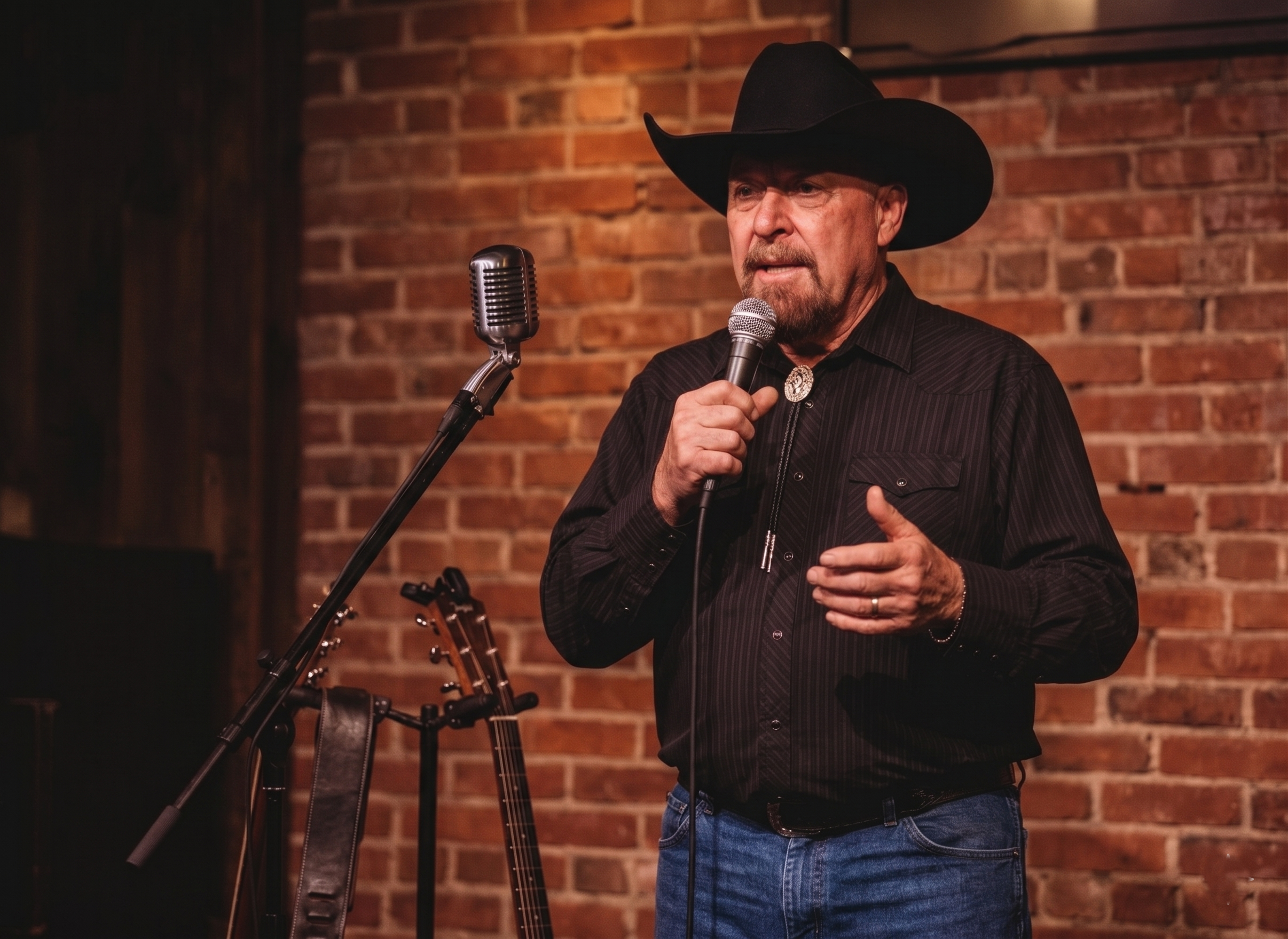 A man wearing a black cowboy hat and black shirt with a bolo tie, holding a microphone, standing in front of a brick wall, with a guitar on a stand nearby.