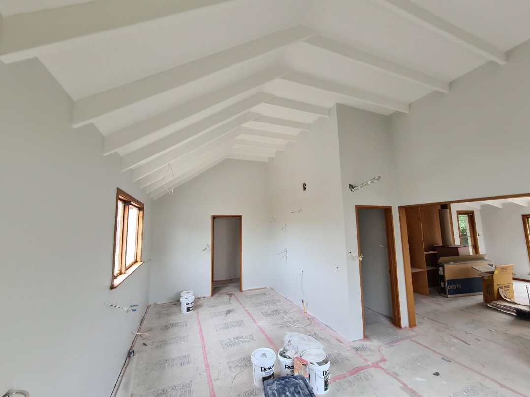 Interior of a house under construction with white walls, a vaulted ceiling with exposed beams, and unfinished floors. There are multiple buckets of paint or construction supplies on the floor and a partially installed TV box and boxes near a built-in wooden shelving unit.