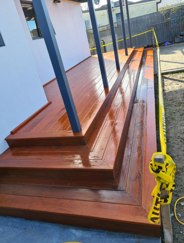 Recently stained wooden deck with steps, metal posts, and a white building wall in the background, surrounded by caution tape.