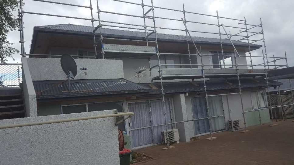 A house under construction or renovation with scaffolding around the upper level, satellite dishes on the roof, air conditioning units outside, and cloudy sky overhead.