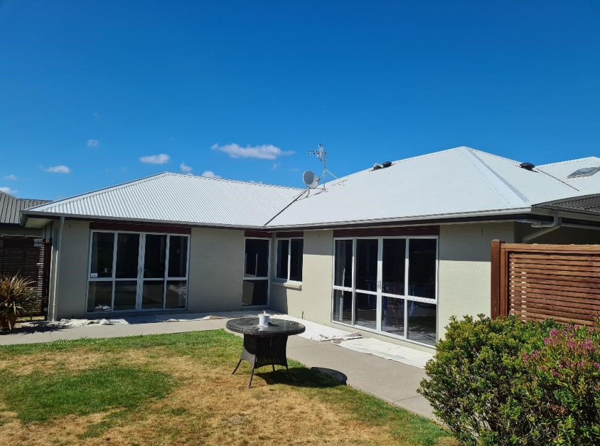 A single-story house with white exterior walls and a white metal roof, set in a garden with a lawn, a concrete pathway, and bushes. The house has large sliding glass doors and windows, and a small round outdoor table in the yard. Clear blue sky with a few scattered clouds is overhead.