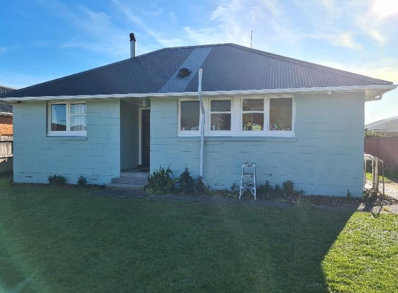 A single-story house with light blue exterior walls, a dark gray metal roof, white-framed windows, a small garden, and a metal ladder in the front yard.