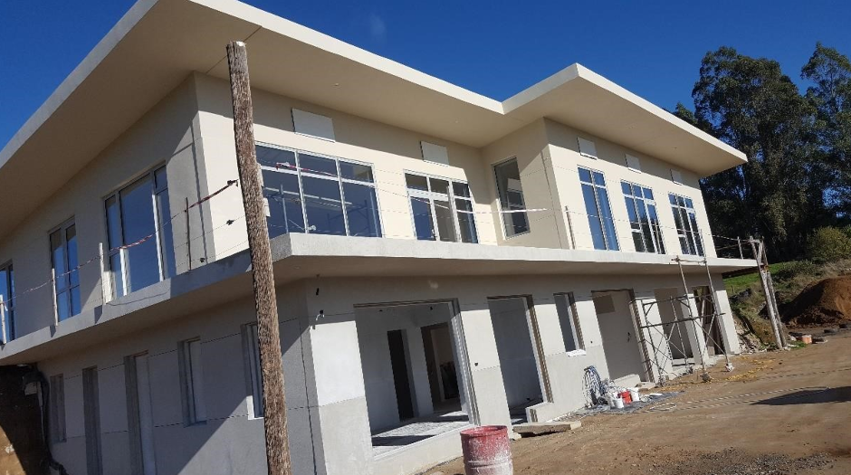 Modern two-story house under construction with large windows, balcony, and scaffolding, on a dirt lot with trees in the background.