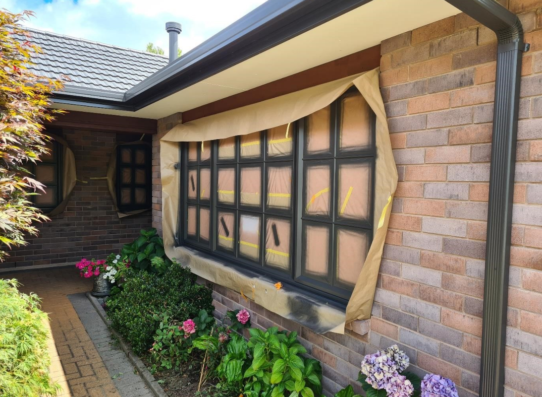 Window under renovation covered with painter's paper taped around edges, on a brick house exterior with flower garden below.