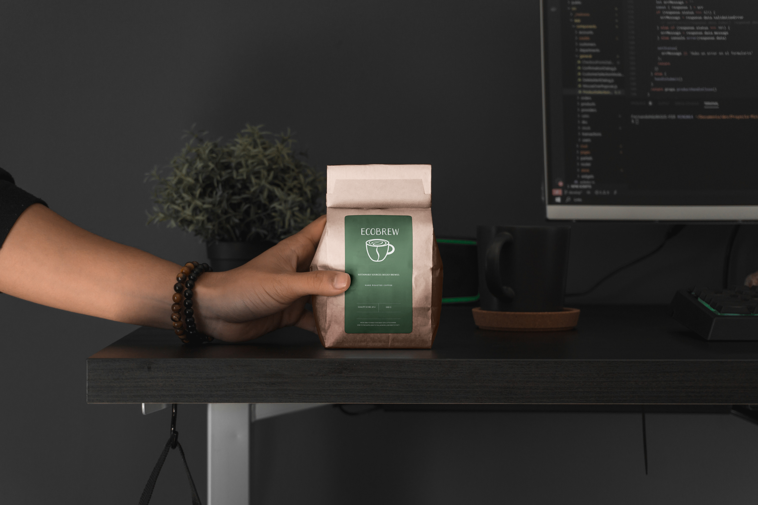 Person holding a brown paper bag of coffee labeled 'ECOBREW' on a dark desk with a plant, a computer monitor displaying code, a black mug, and a keyboard in the background.