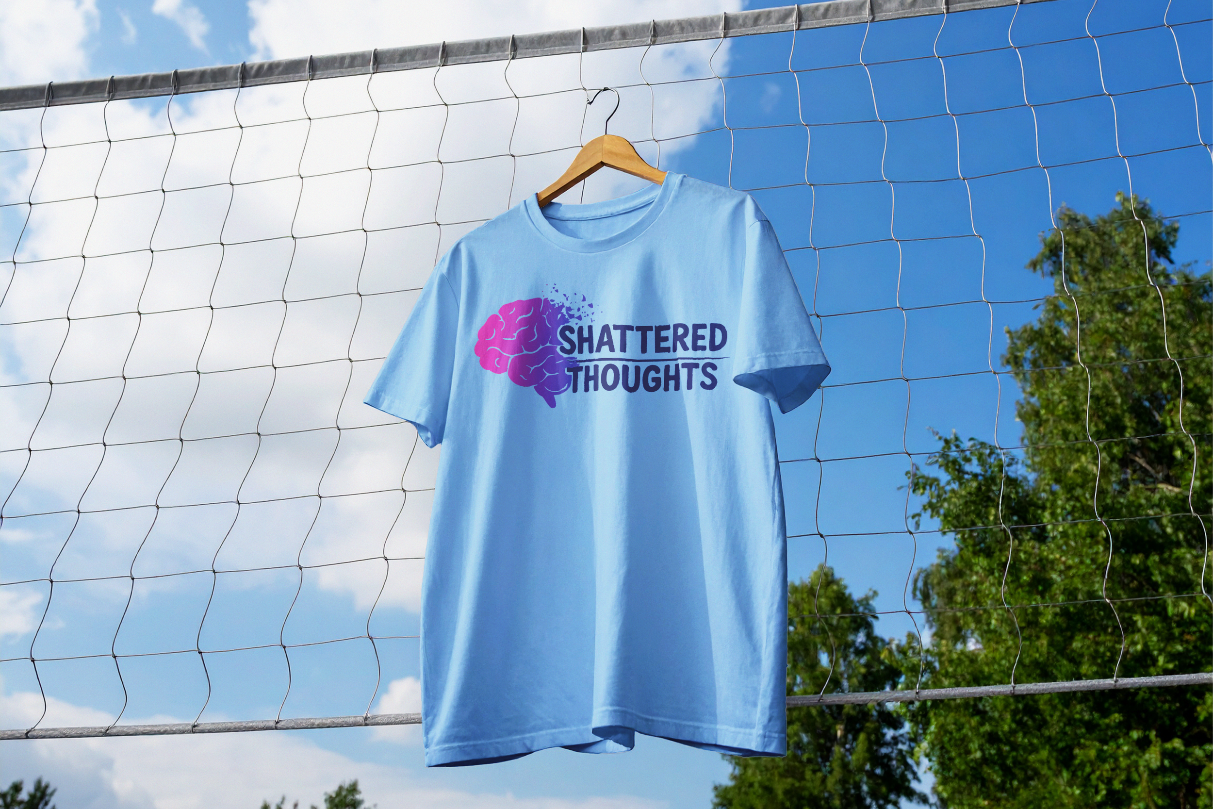 A blue T-shirt hanging on a wooden hanger on a wire fence outdoors, with trees and a blue sky with clouds in the background. The T-shirt has a purple and pink graphic of a brain and the words "SHATTERED THOUGHTS" printed on it.