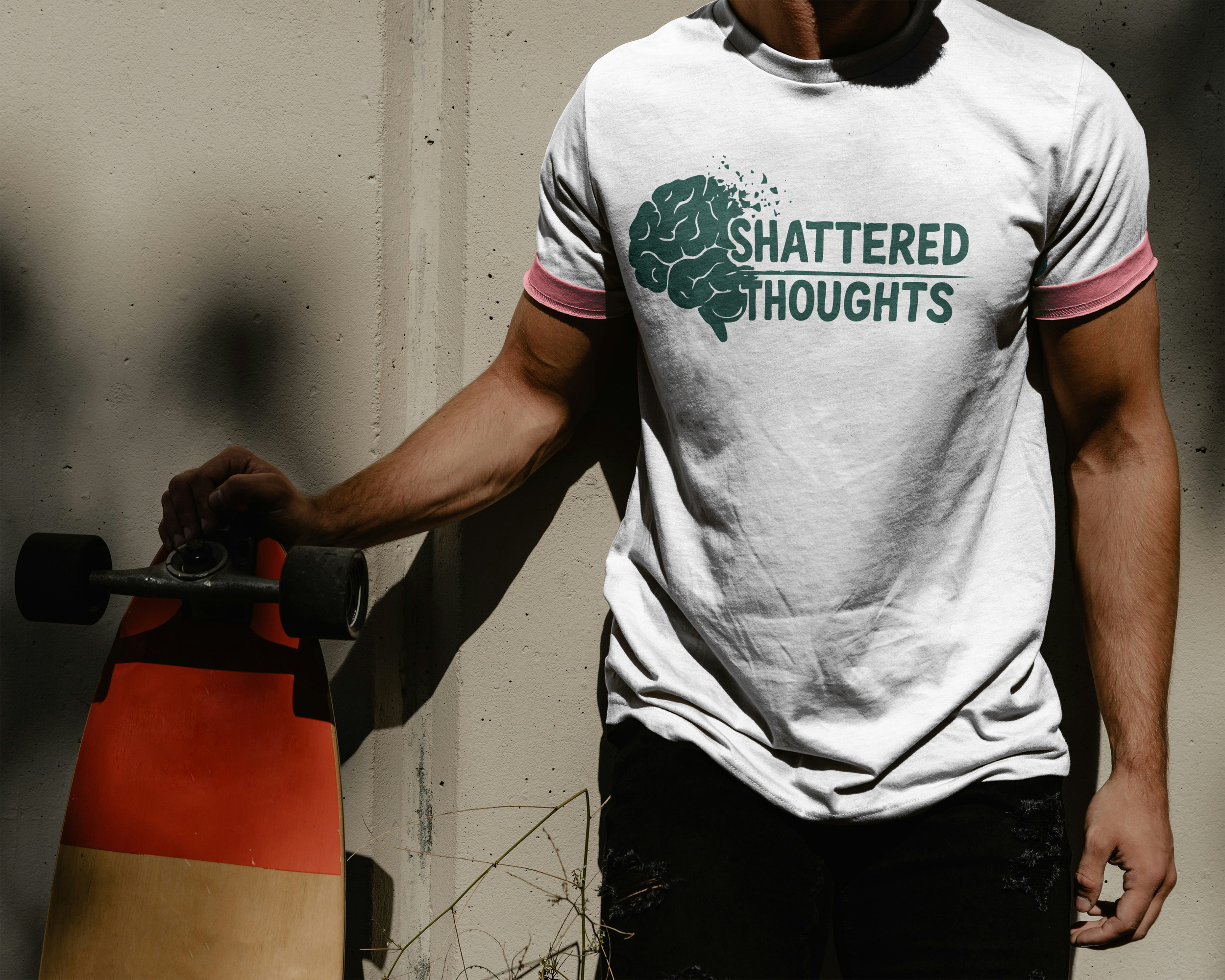 Person with a skateboard standing against a concrete wall, wearing a white T-shirt with a brain and shattered text graphic that reads "Shattered Thoughts".