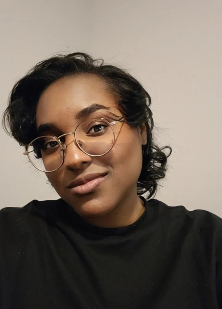 A young woman with short, curly dark hair, wearing round glasses and a black shirt, smiling slightly against a plain gray background.