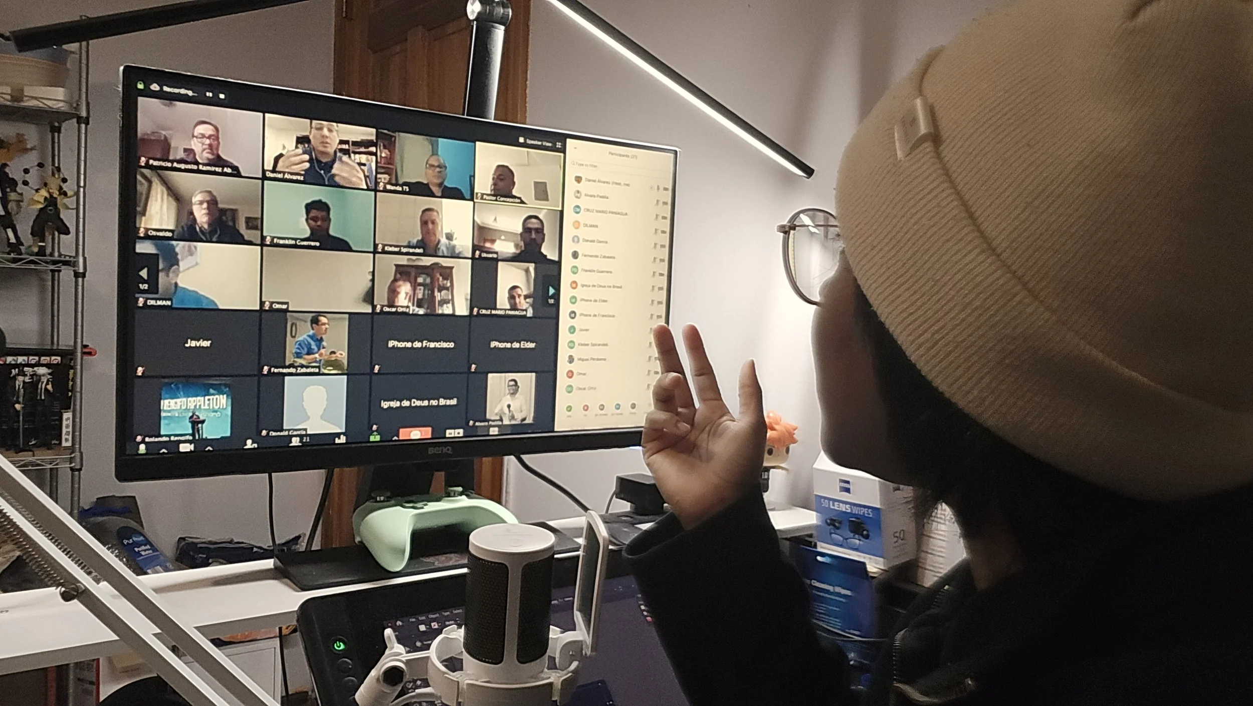 A person wearing a beige beanie making a peace sign in front of a computer monitor displaying a video conference with multiple participants.