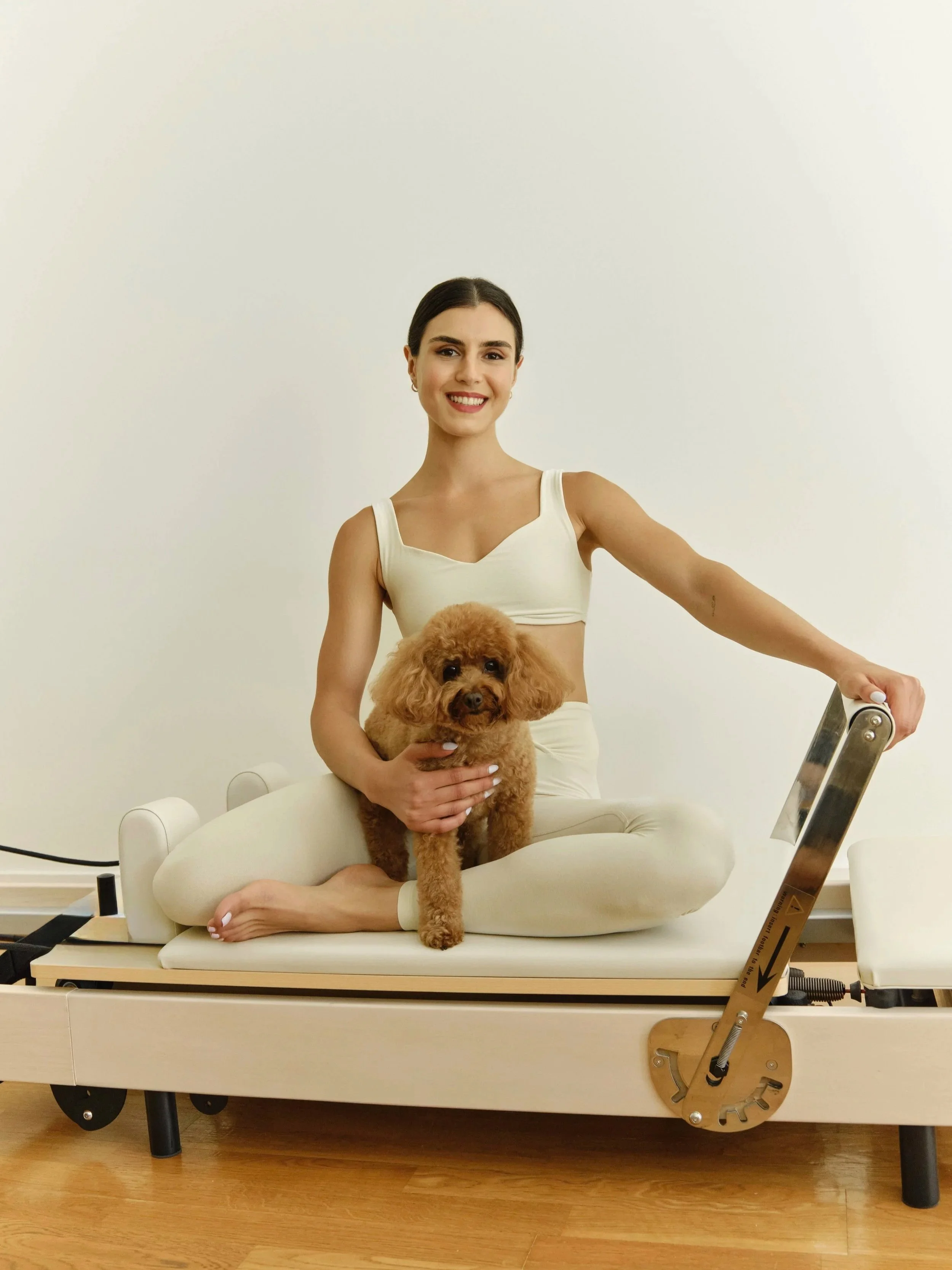 A woman sitting cross-legged on a medical or therapy table with a small brown poodle on her lap, holding it with both hands, in a room with plain white walls.