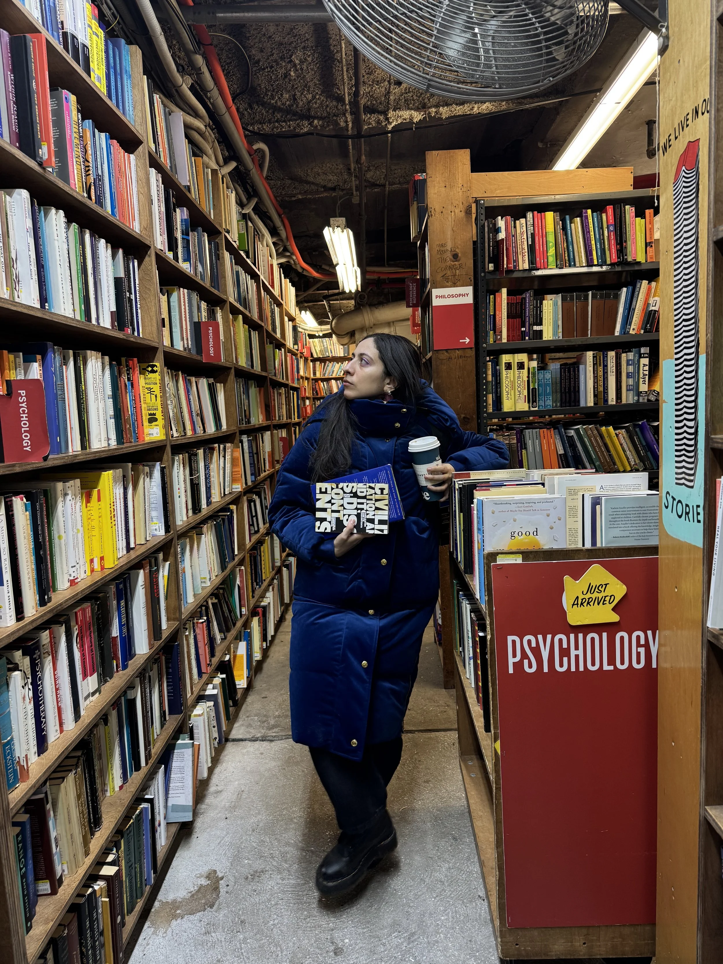 A woman in a blue coat and black boots holding a coffee cup and a book in a bookstore aisle, looking to her left. Bookshelves around her are filled with various books, with signs indicating sections like philosophy and psychology.