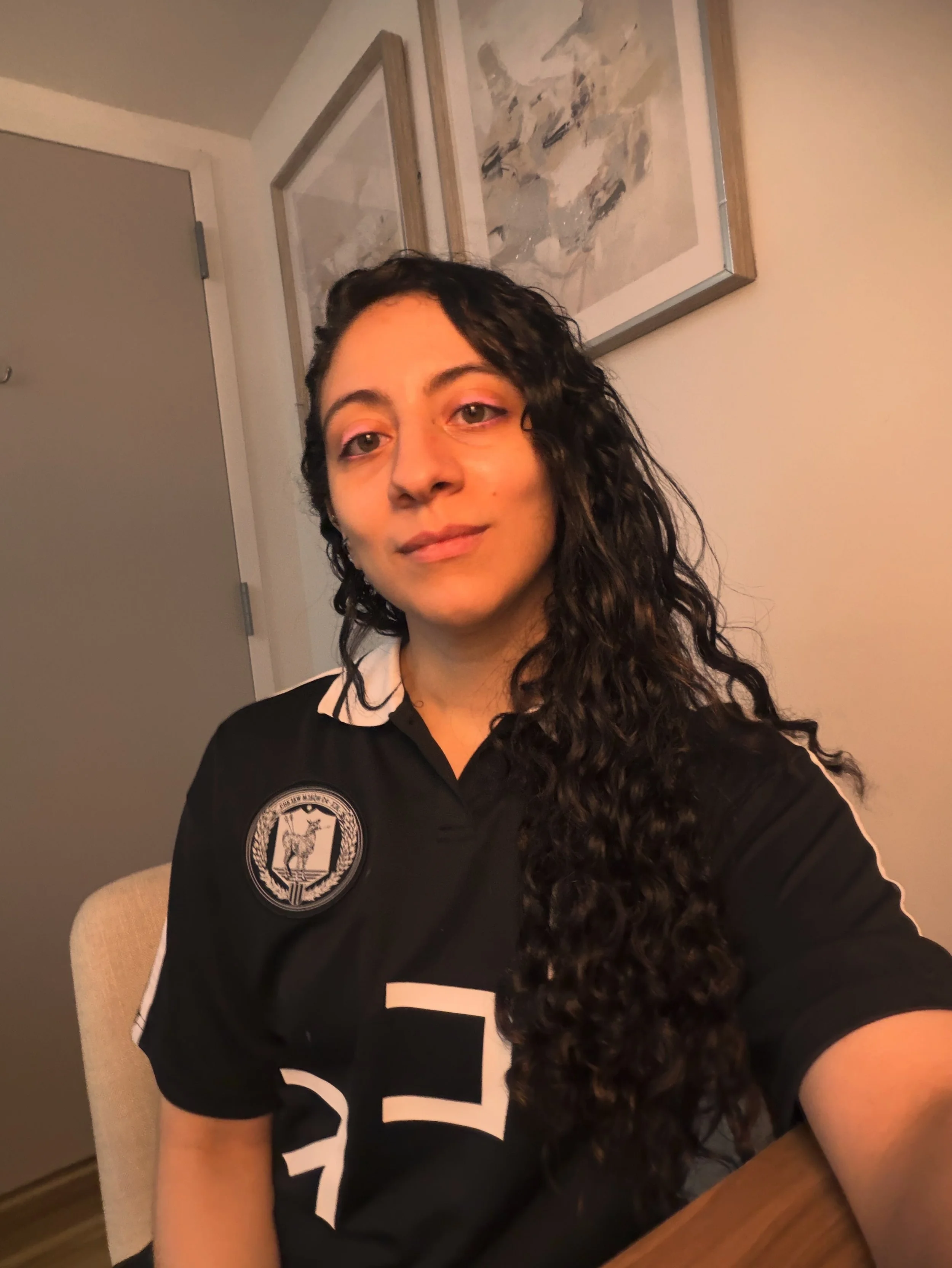 A woman with long curly dark hair is sitting in a room with beige walls and a door. She is wearing a black shirt with white letters and a logo on the sleeve. Behind her, there are two framed abstract paintings hanging on the wall.