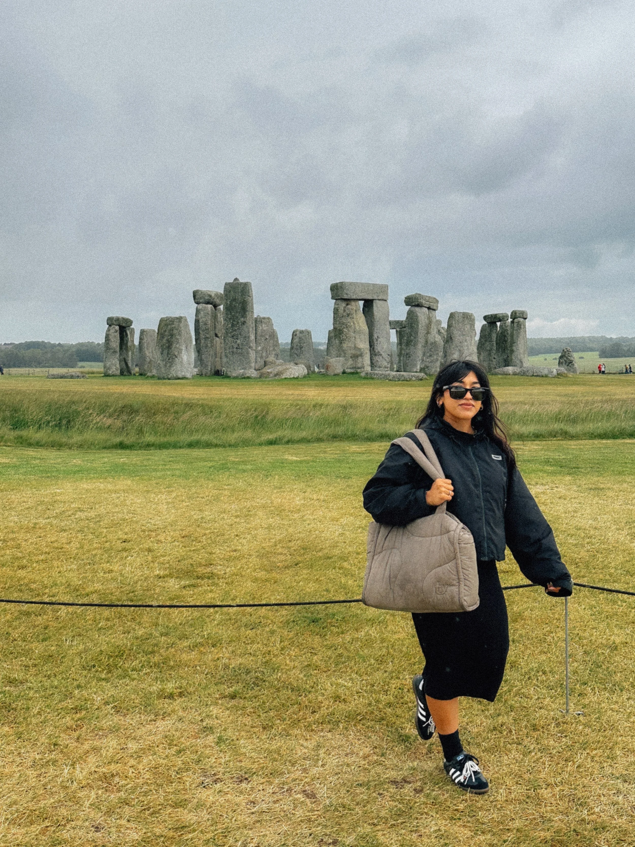 A woman carrying a bag and wearing sunglasses stands in front of Stonehenge on a cloudy day.