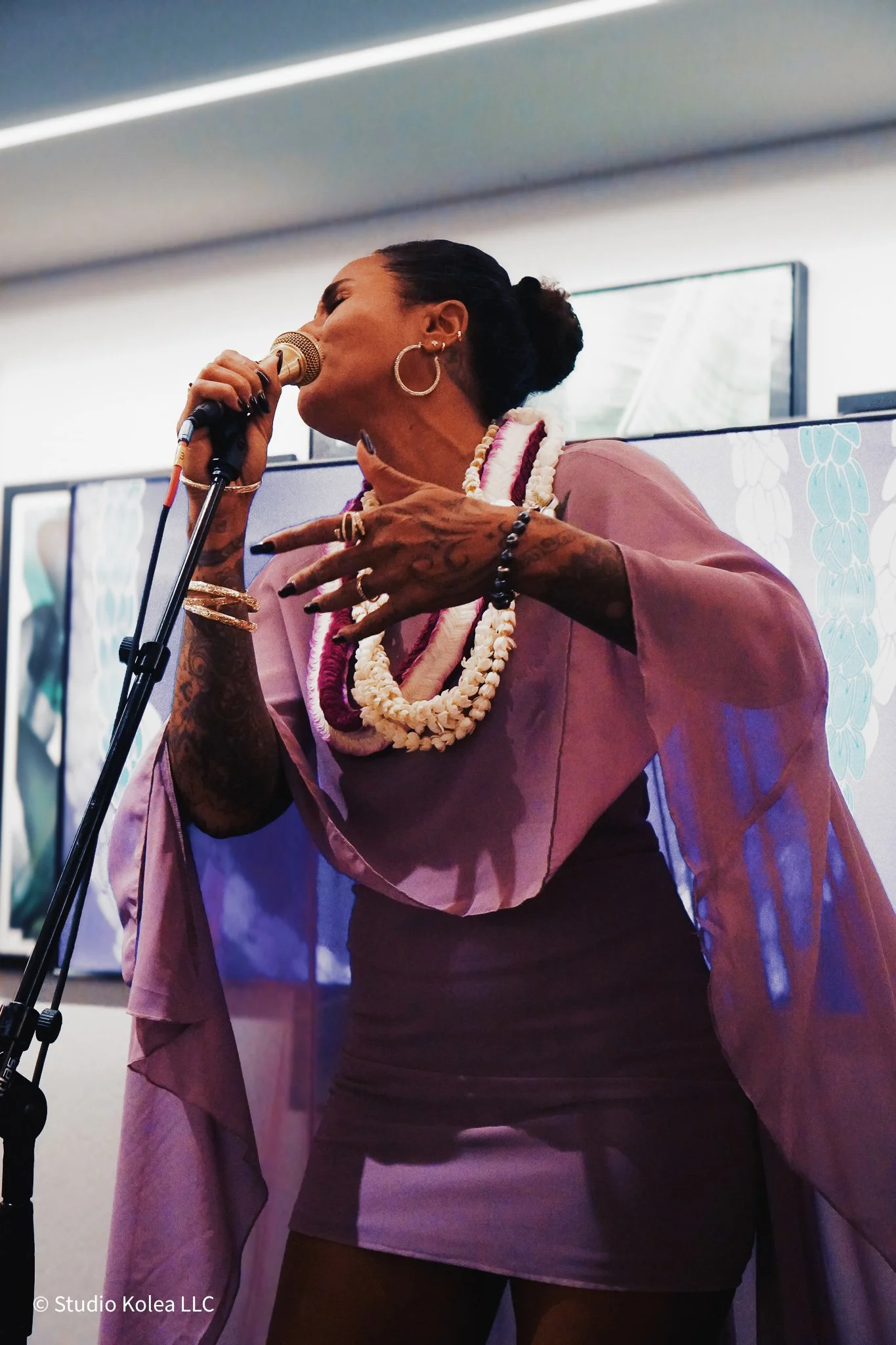 A woman with dark hair tied back, wearing jewelry, singing into a microphone at an art gallery or exhibition.