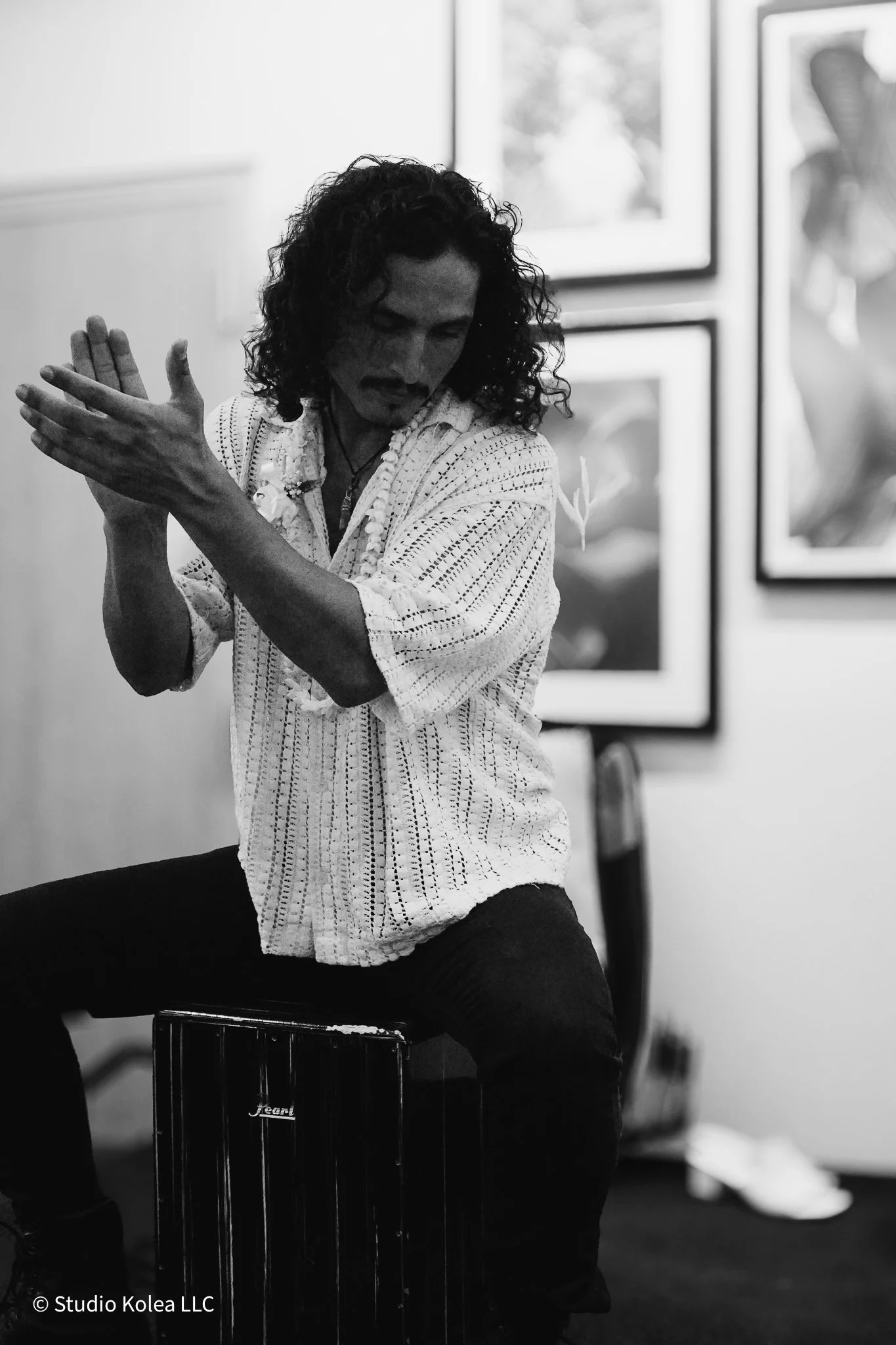 A man with long curly hair wearing a white patterned shirt, sitting on a black drum, posing with his hands extended in a dance or performance studio with framed artwork on the wall behind him.