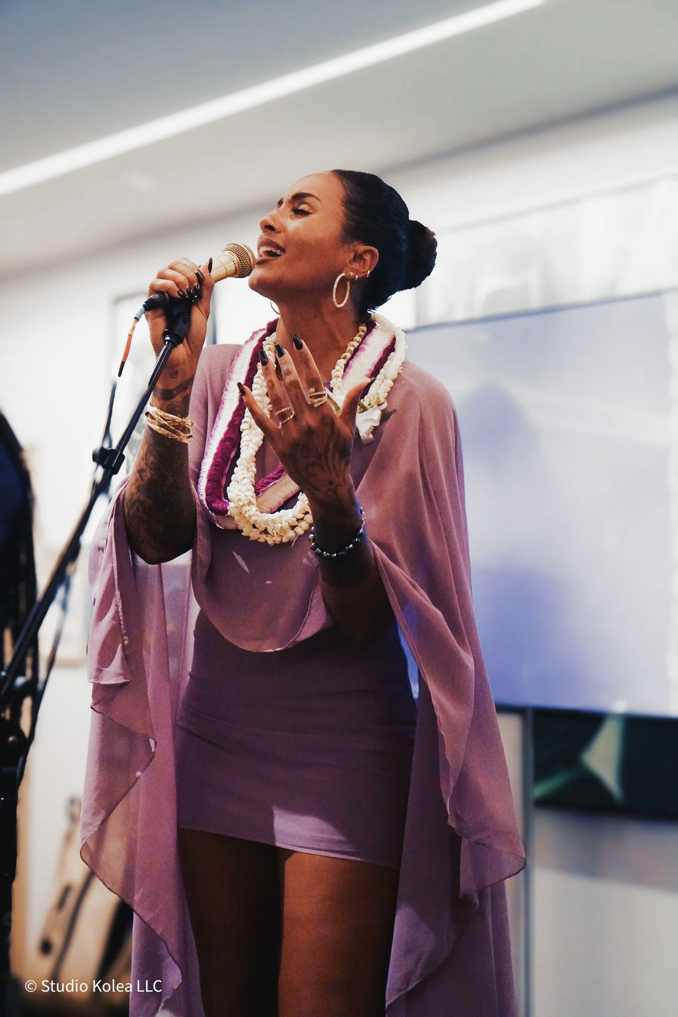 A woman with dark hair tied back, wearing hoop earrings, multiple necklaces, bracelets, and rings, singing into a microphone on stage with a large display screen in the background.