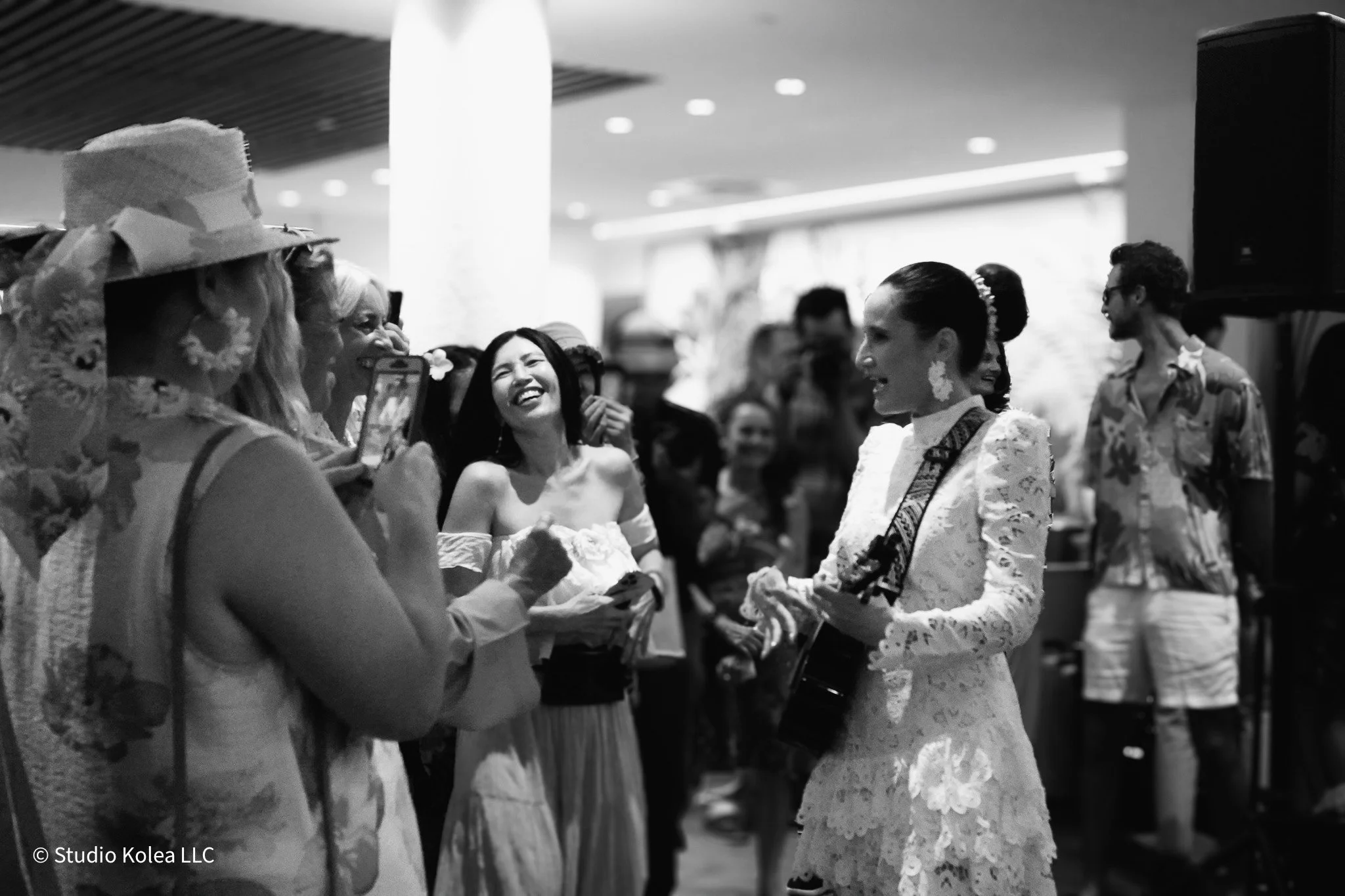 A woman playing guitar and singing at a social gathering, surrounded by people enjoying the moment with smiles and laughter in an indoor venue.
